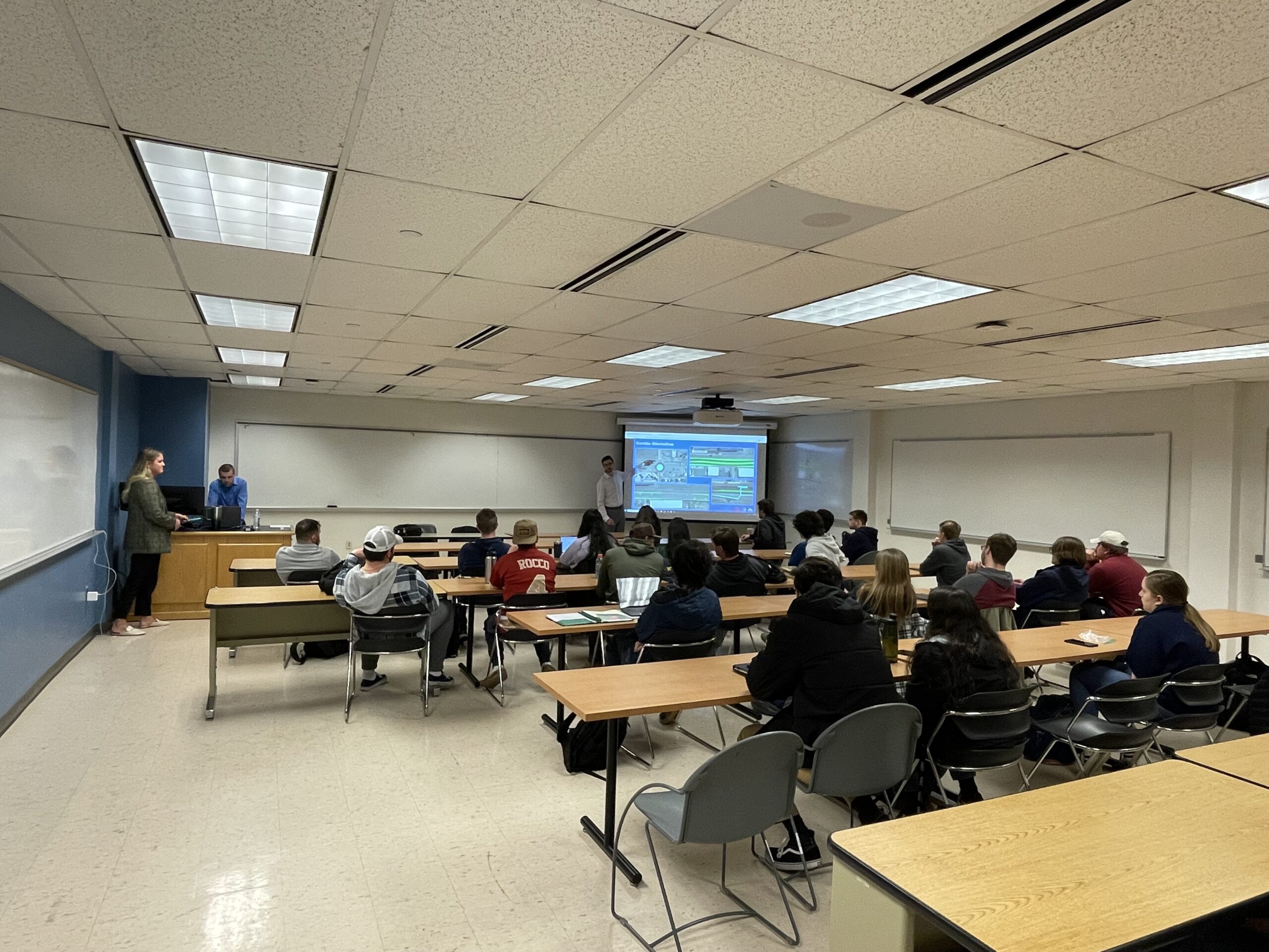 TCNJ students sitting in a classroom (backs to the camera) listening to the two presenters in front of them. 