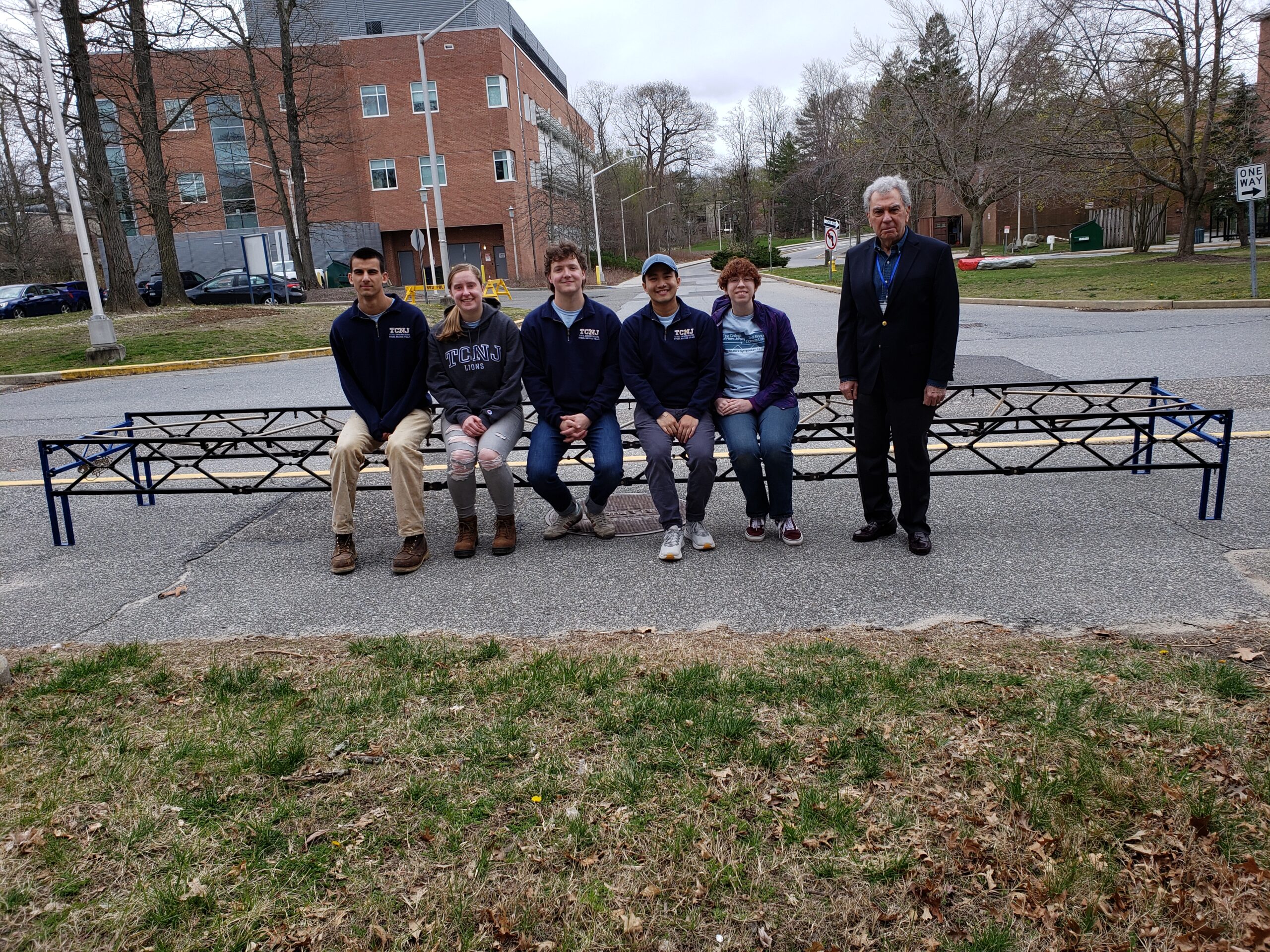 Five students sitting on a completed steel bridge. Faculty advisor is standing next to them. Photo was taken outside the competition venue. Academic buildings in the background.