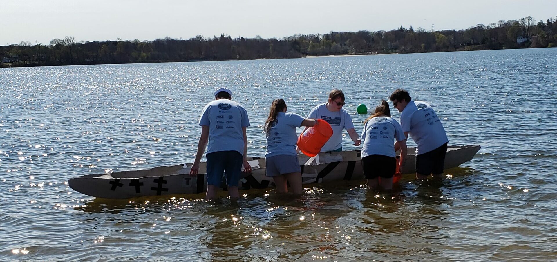 TCNJ students standing in a lake around a canoe holding orange buckets and filling the canoe with water.