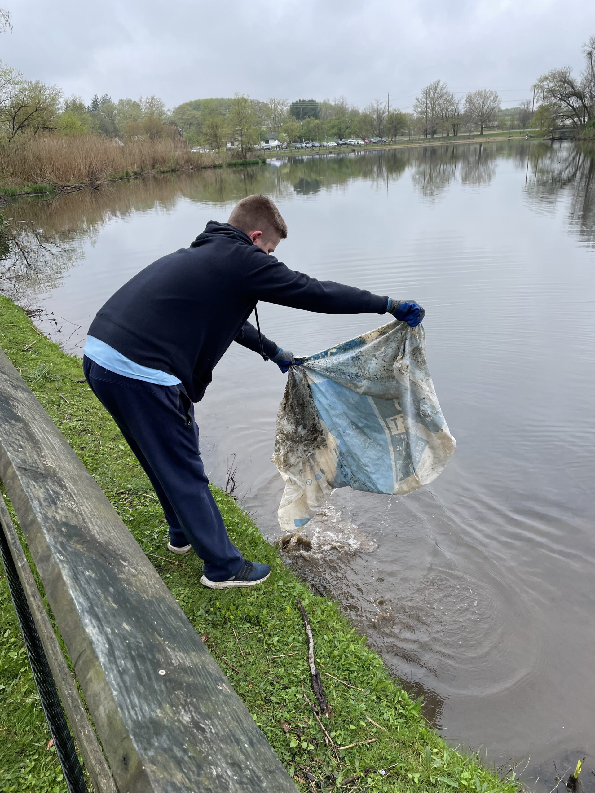 A student standing at the edge of a lake pulling a large plastic bag out of it.