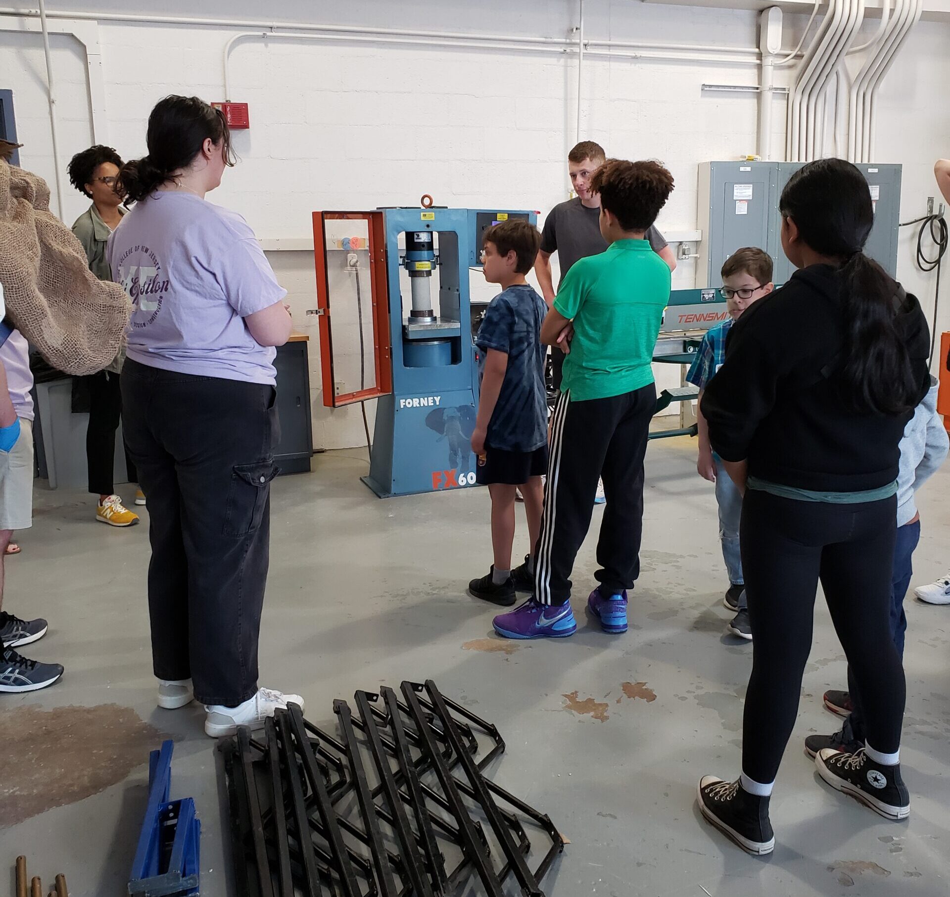 A group of children standing in front of a compressive texting machine observing a compressive test on a concrete cylinder.