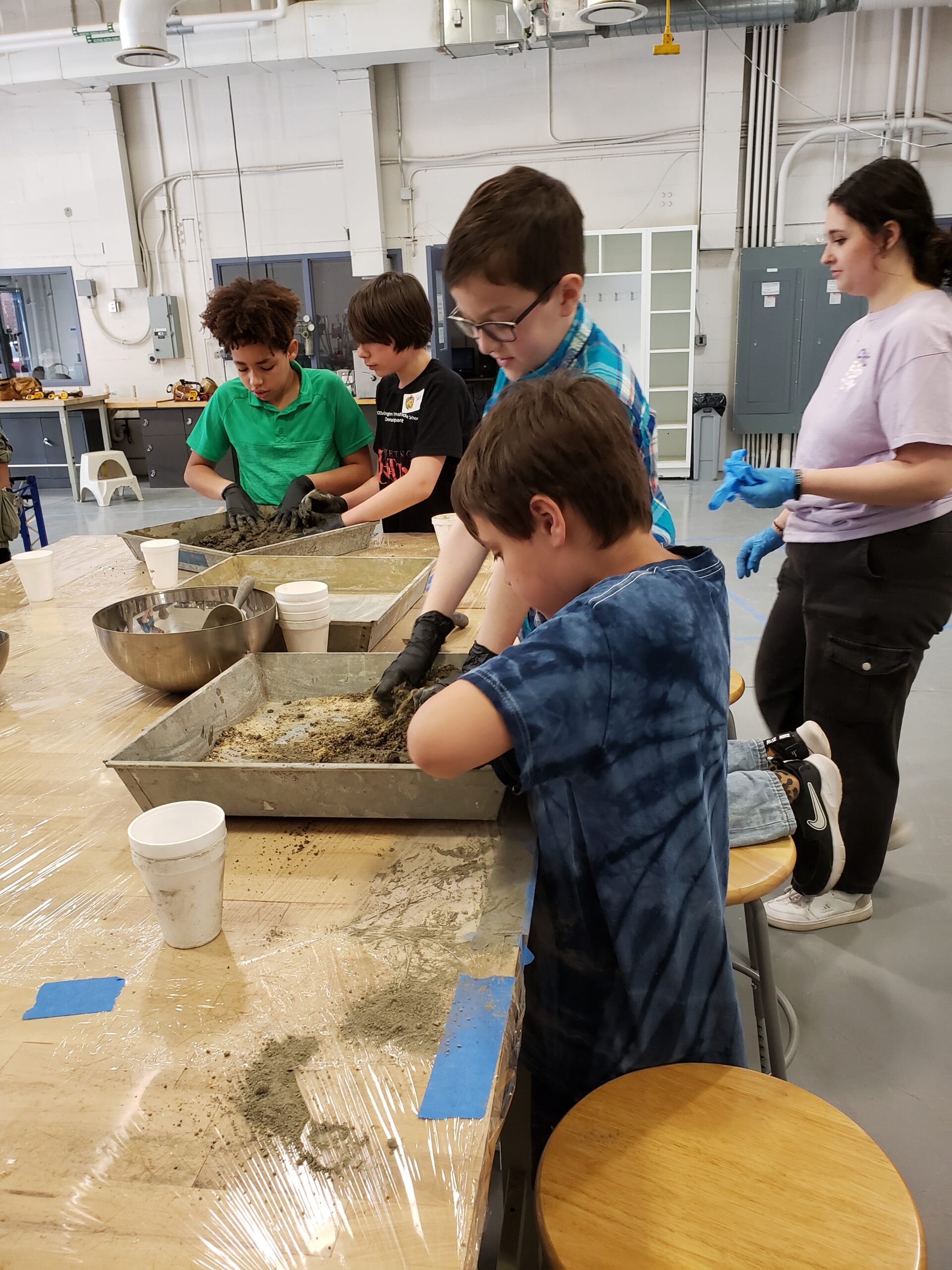A group of children stands around a table mixing concrete inside large pans.