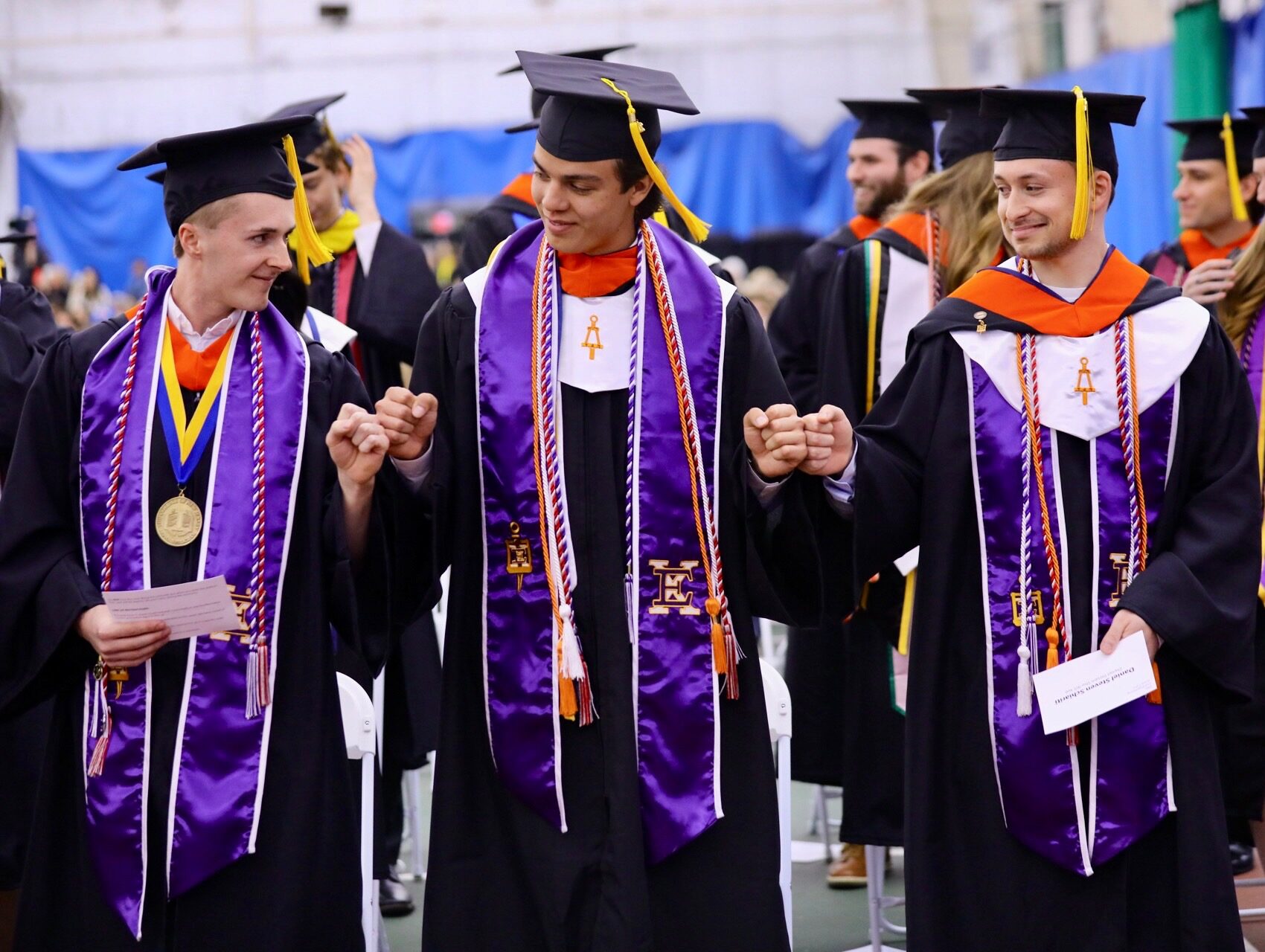 Three students in cap and gowns stand and fist bump each other.