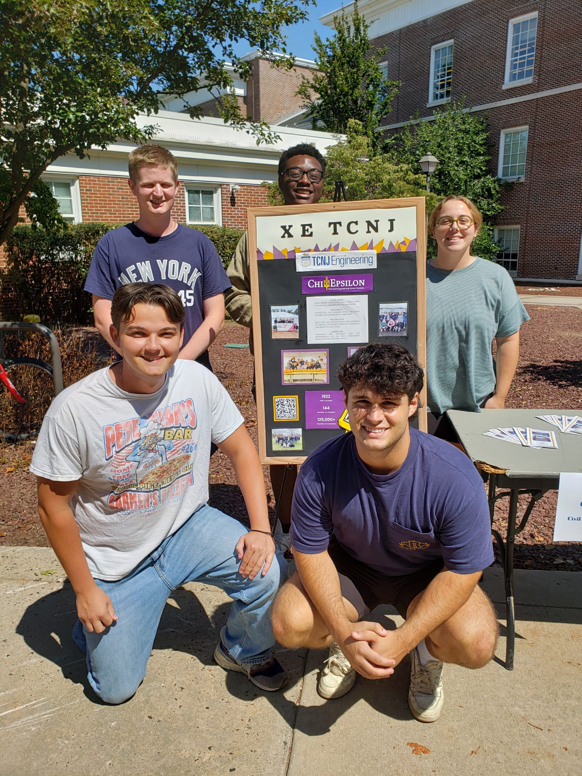 Three students stand around, and two students kneel in front of a board that represents TCNJ XE Chapter information.