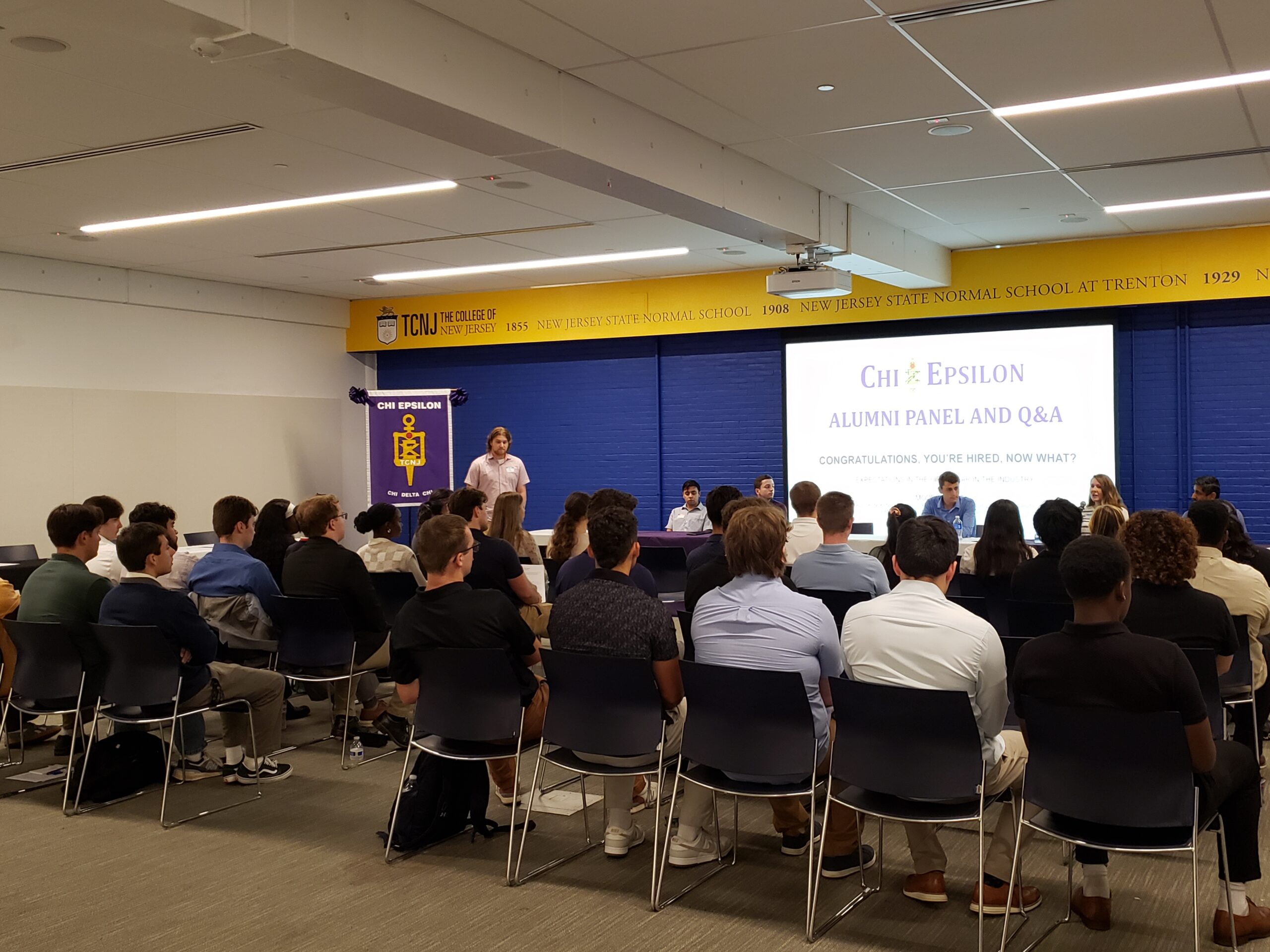 A large group of students was photographed from the back. Students are sitting in front of a panel of alumni with a screen behind them.