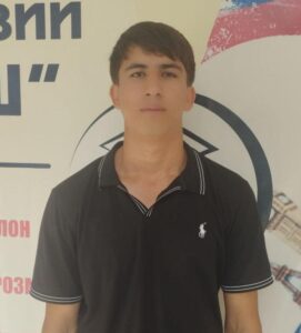 A young man with dark hair in black polo shirt stands in front of a wall.