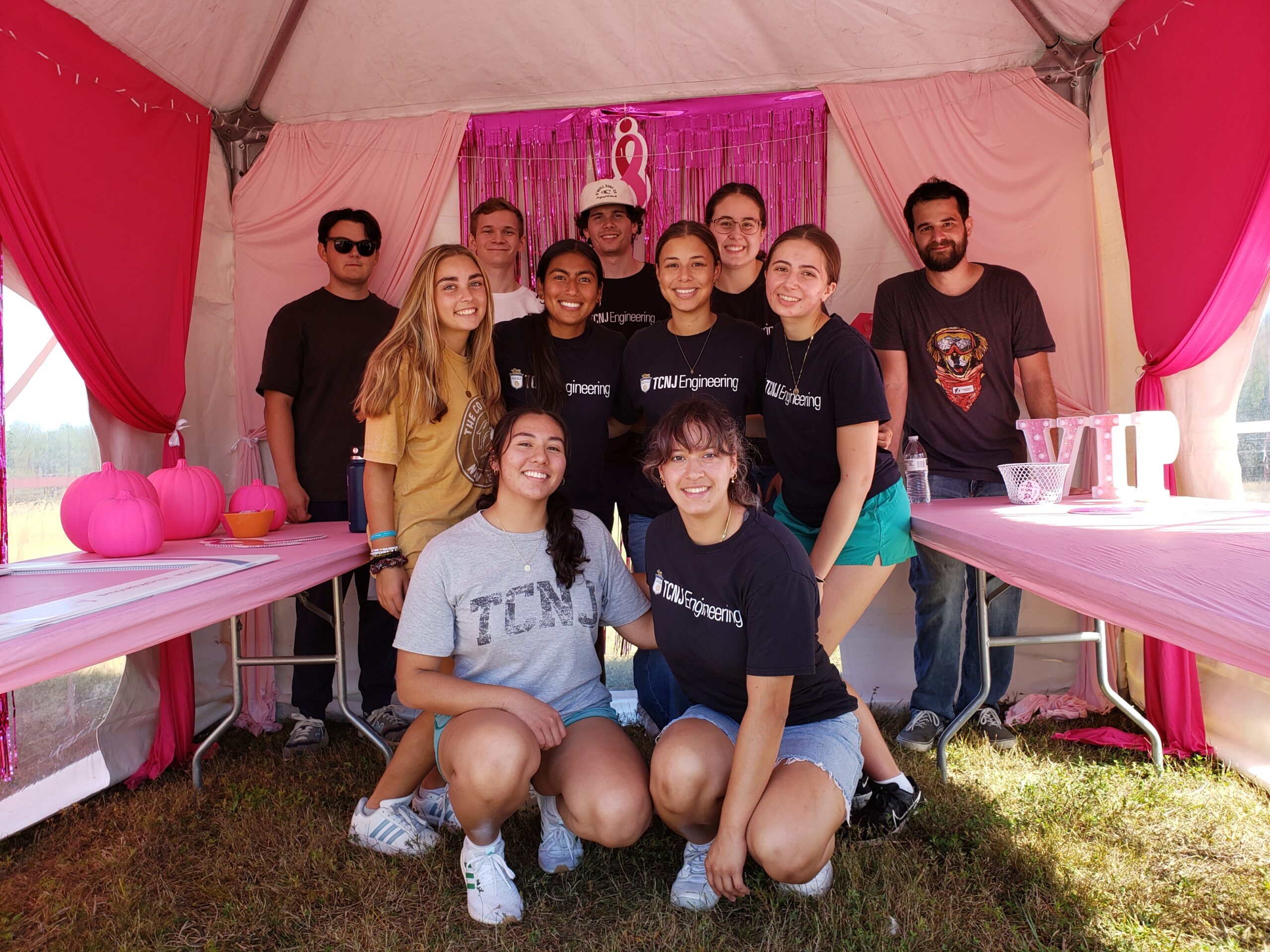 A group of 11 students stands inside a tent with a backdrop of pink curtains.