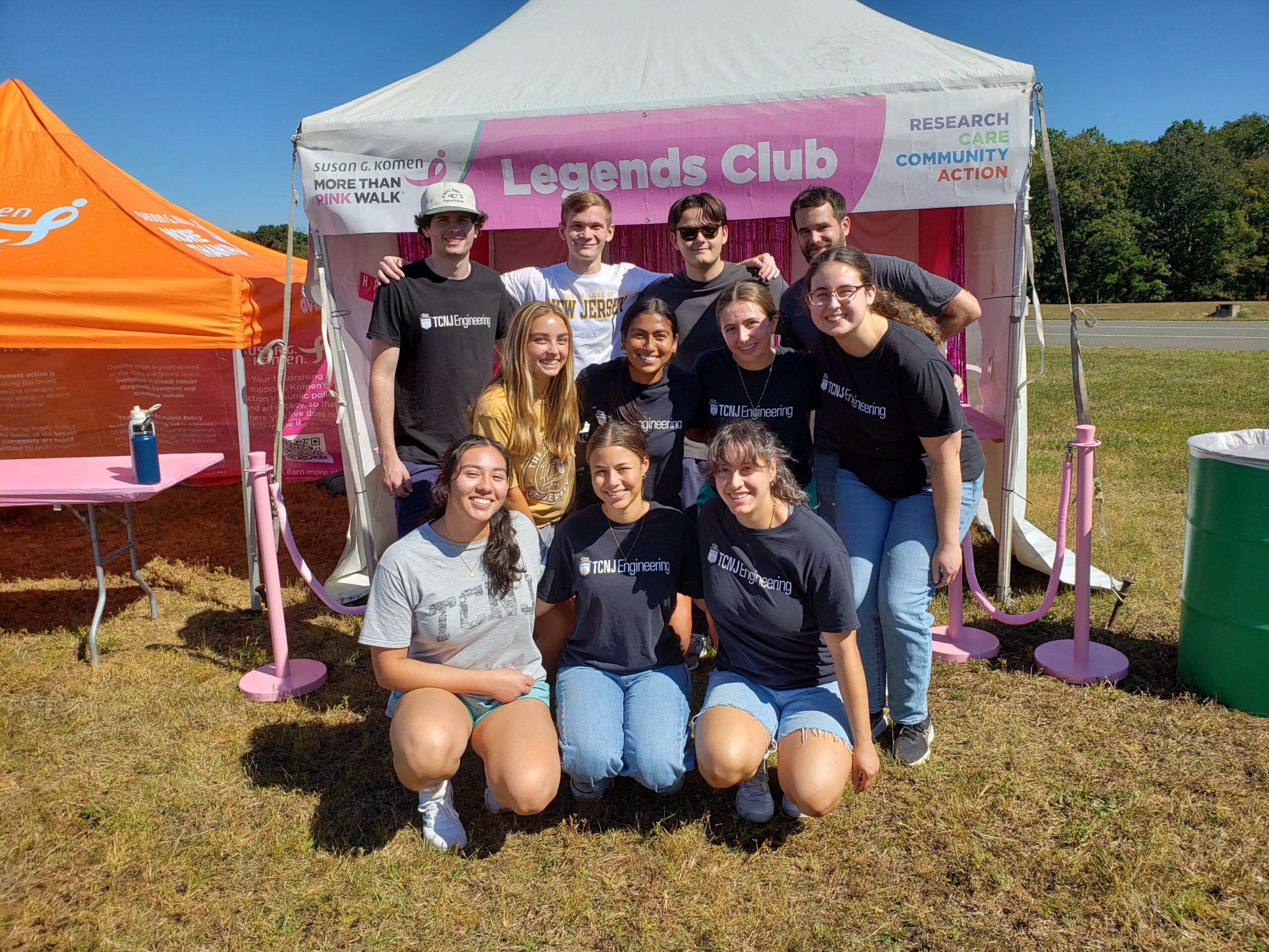 A group of 11 students stands at the entrance to a small tent that displays the Susan G. Komen logo and the words Legends Club.