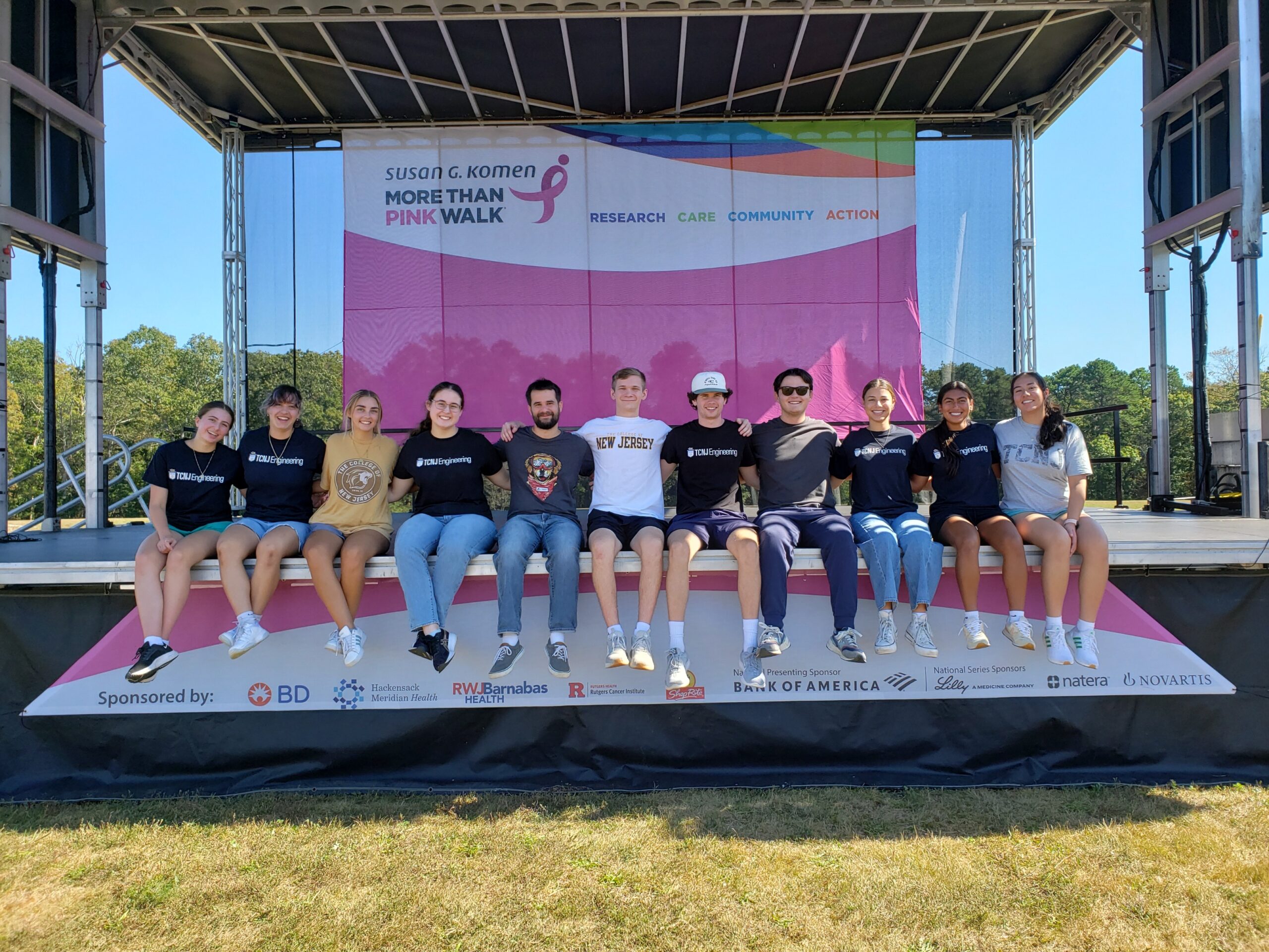 A group of 11 students sits at the edge of a stage. Backdrop is a Susan G. Komen banner.