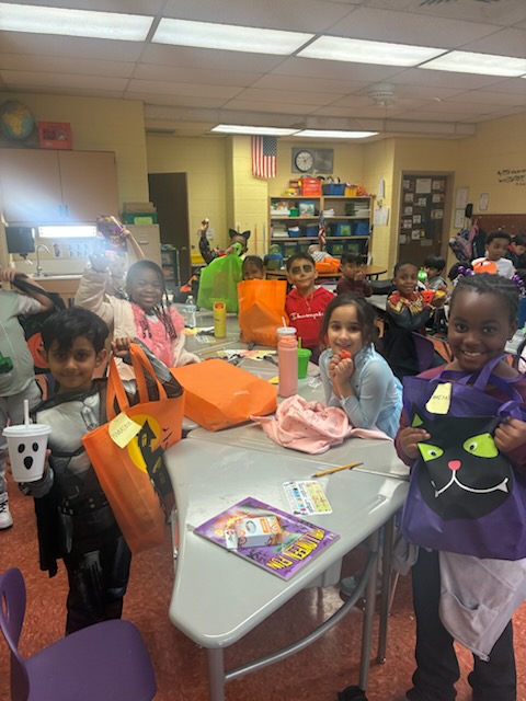 A group of first grade students in Halloween costumes sit at a large table looking at Halloween bags, toys and candy.