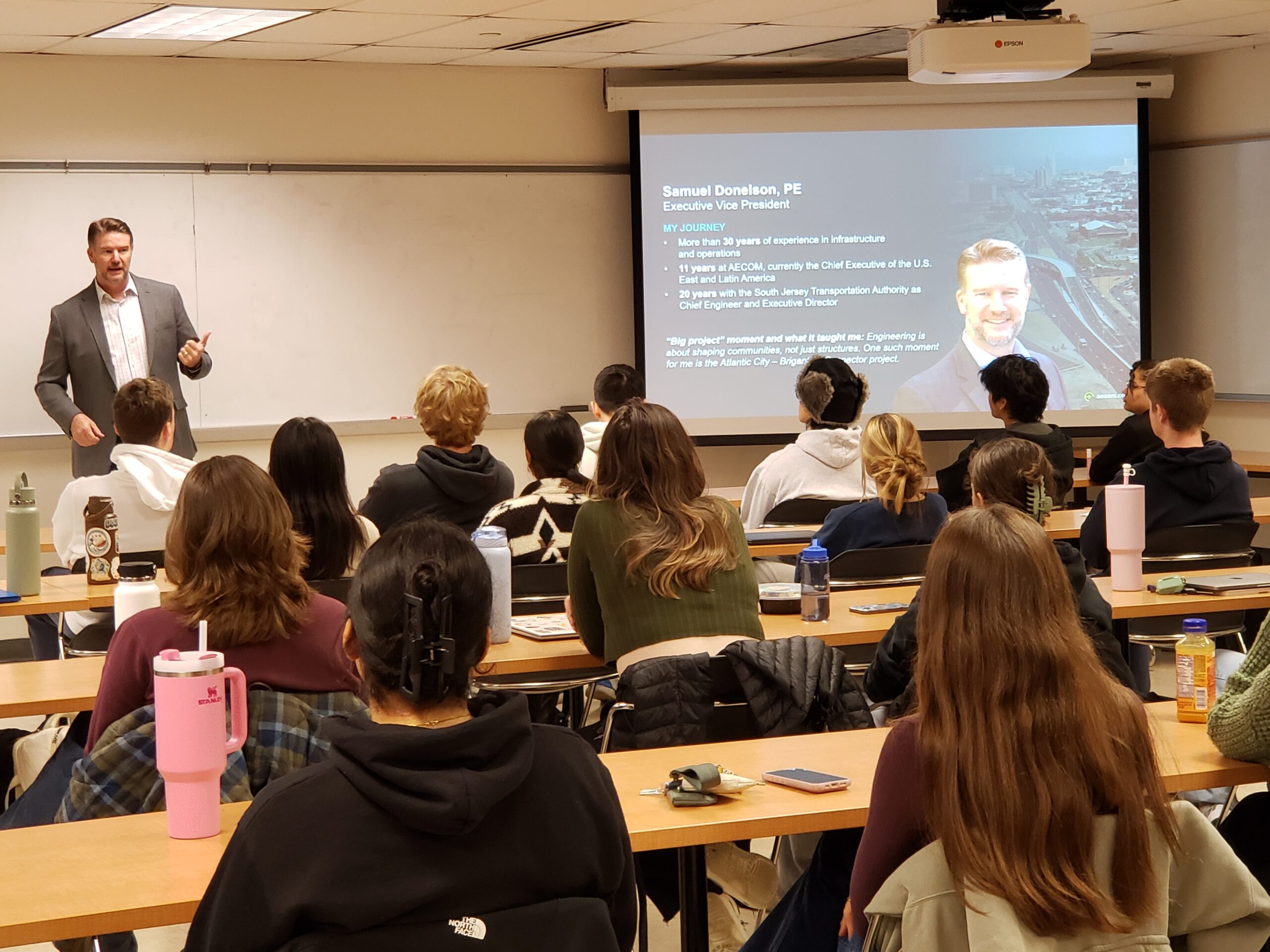 A male presenter stands next to a screen in front of the room full of students.
