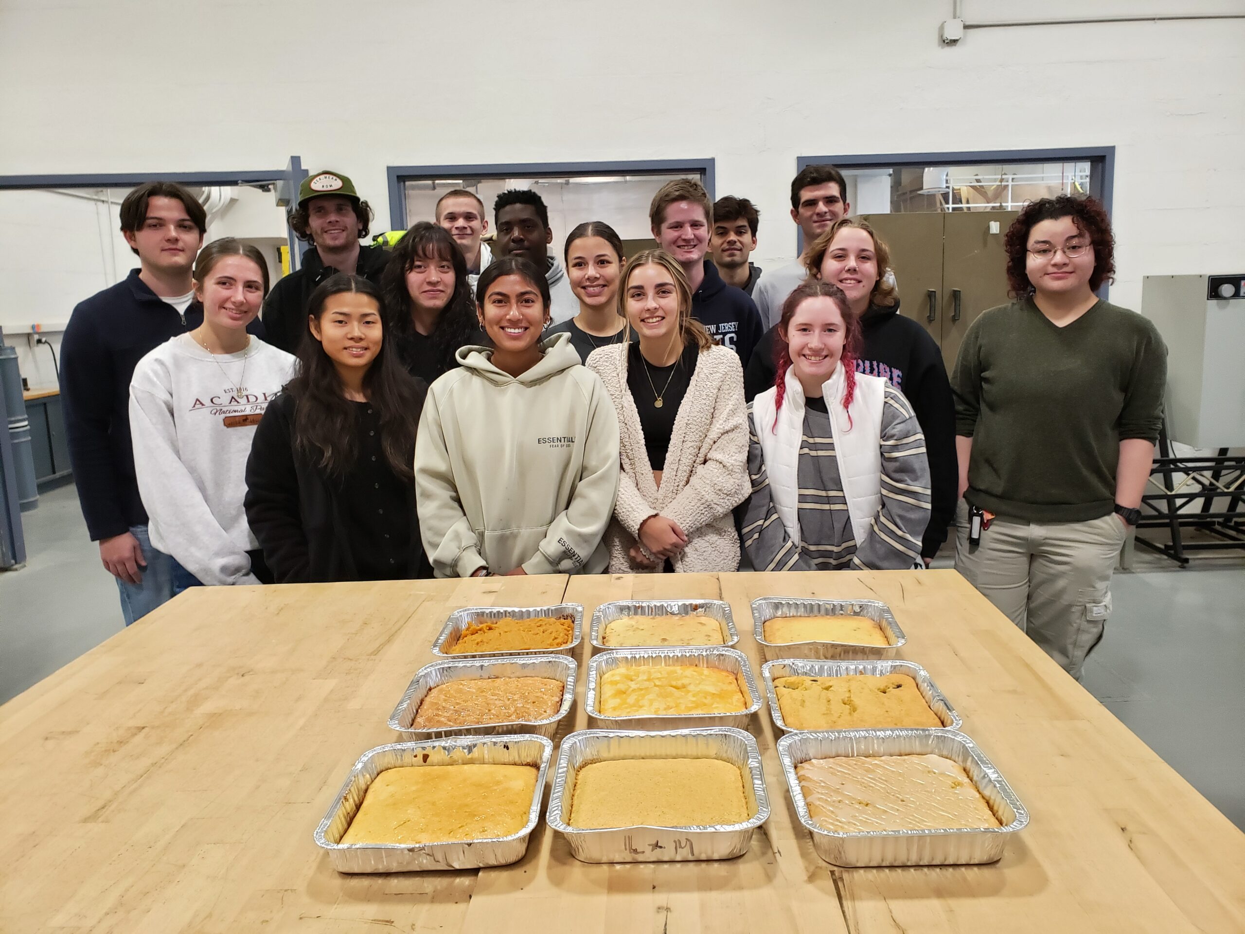 A group of students stand behind a table. There are nine square pans with cornbread on the table.