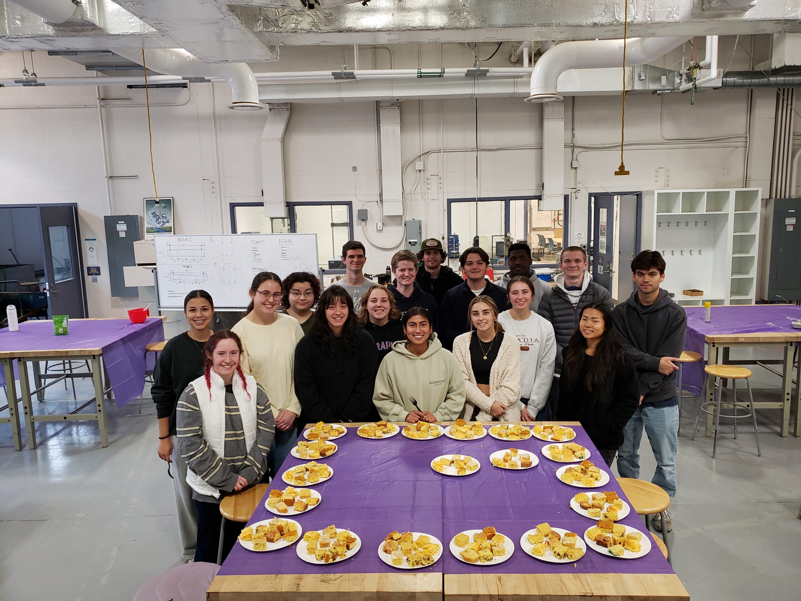 A group of students stands around a table. There are multiple plates on the table, each having small pieces of cornbread.