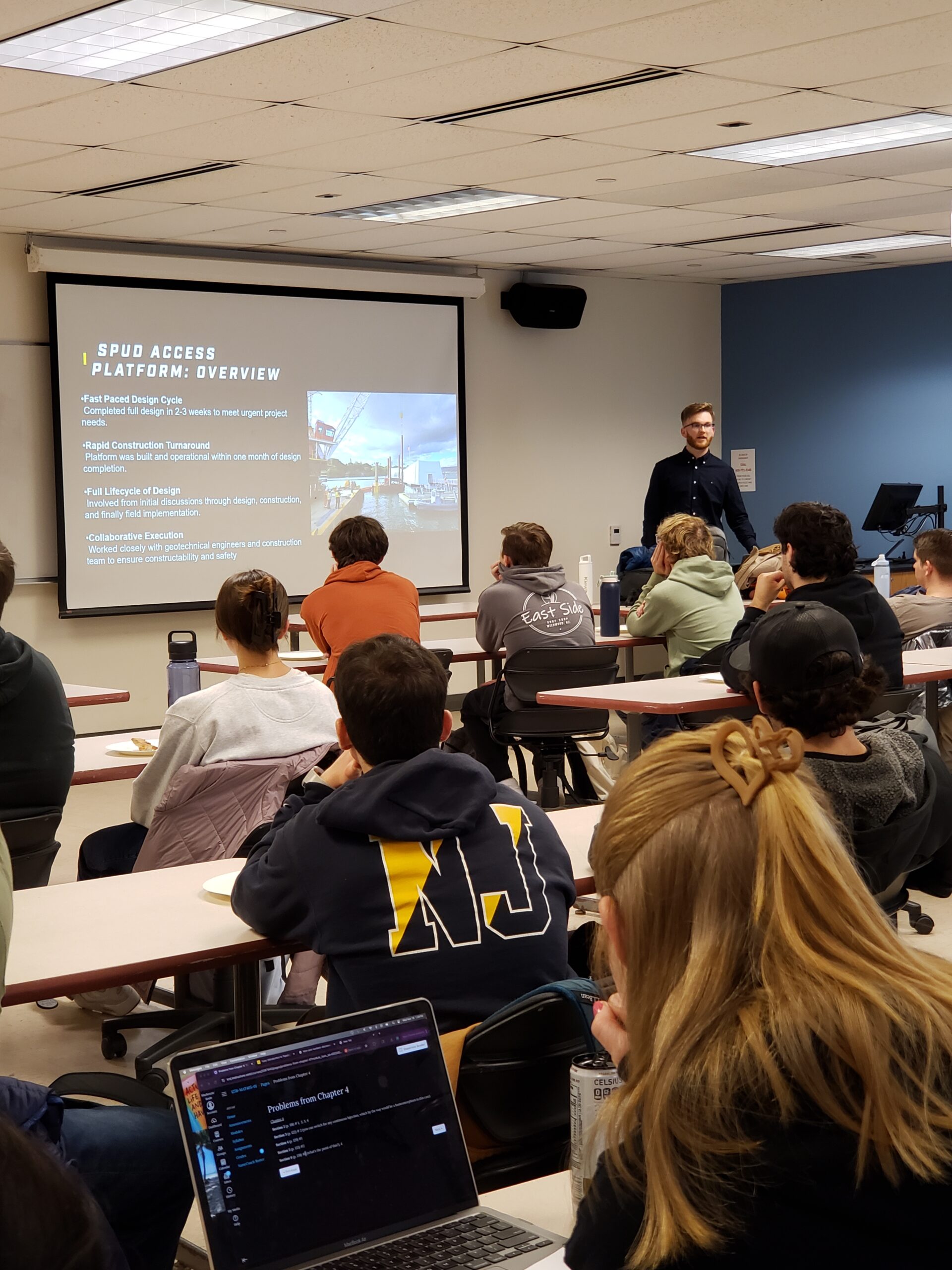 A presenter stands to the right of a screen in front of a room of students who are sitting at their tables.