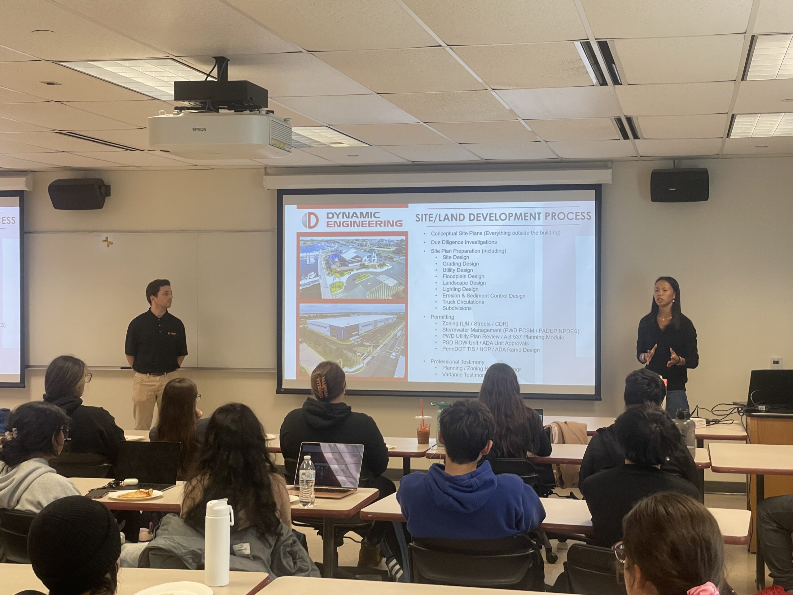 A young male and a young female professional stand on each side of a screen while talking to a room full of students.