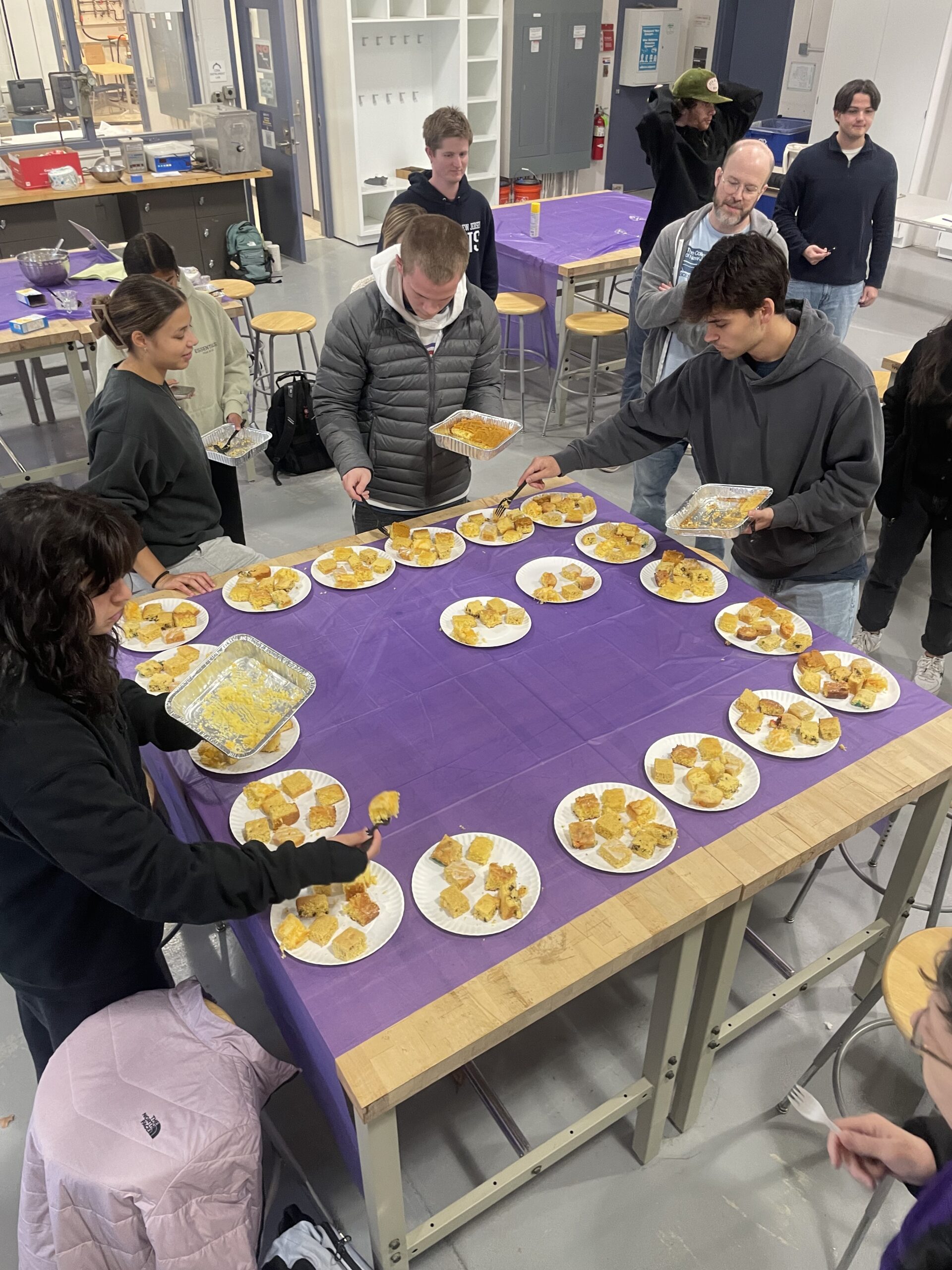 Students placing cut cornbread pieces on plates arranged around the edge of the table