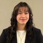 A headshot of a female student with long dark brown hair in a black suit and white shirt with a gold cross necklace.