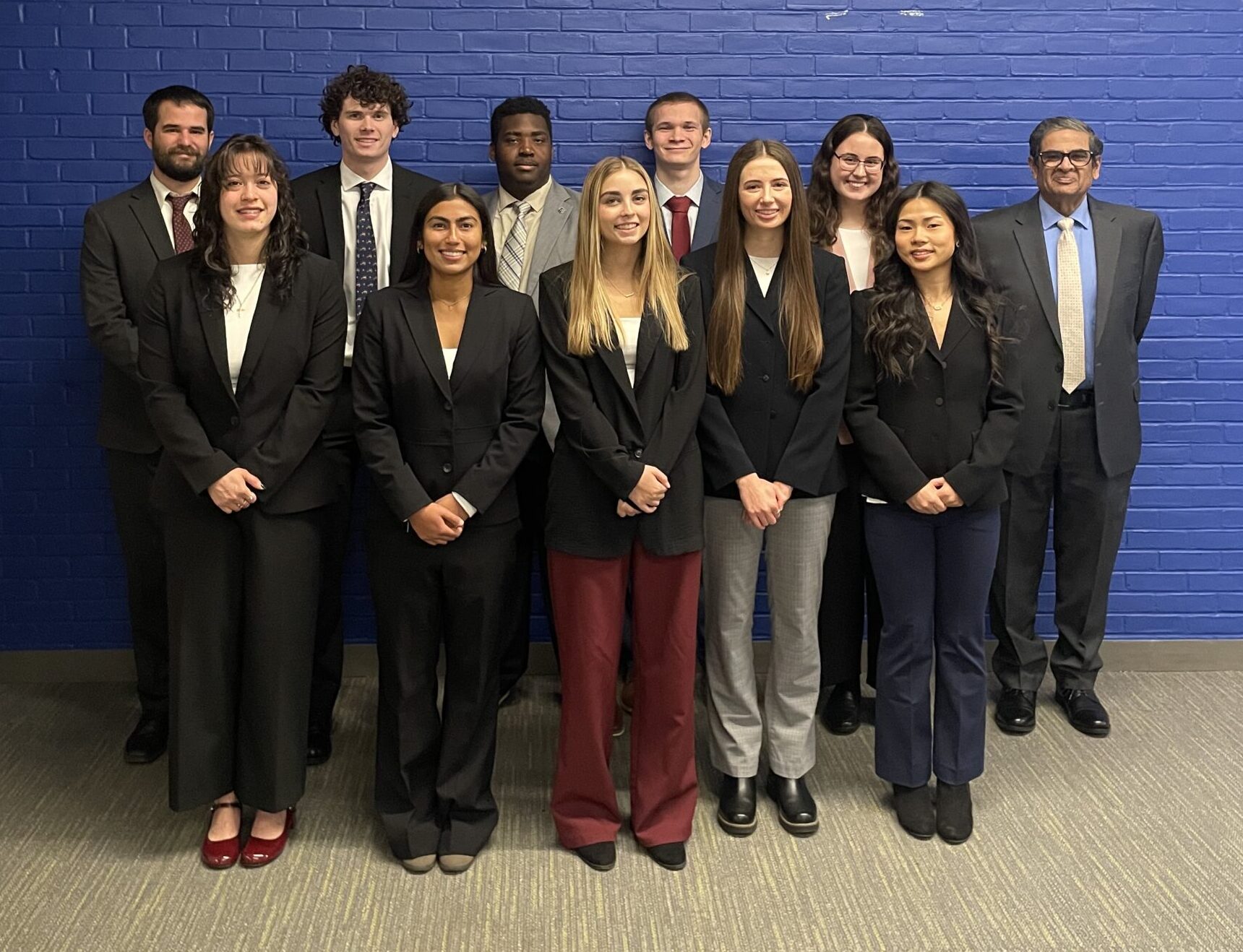 A group of people in business suits stands in front of a blue brick wall.
