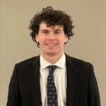 A headshot of a male student with curly brown hair in a dark suit, white shirt and a dark patterned tie