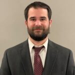 A headshot of a young male with beard, white shirt and red tie
