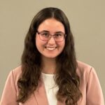 A headshot of a female students with long brown hair and glasses in a pink suit and white shirt
