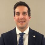 A headshot of a male in a dark suit, dark tie, and a white shirt