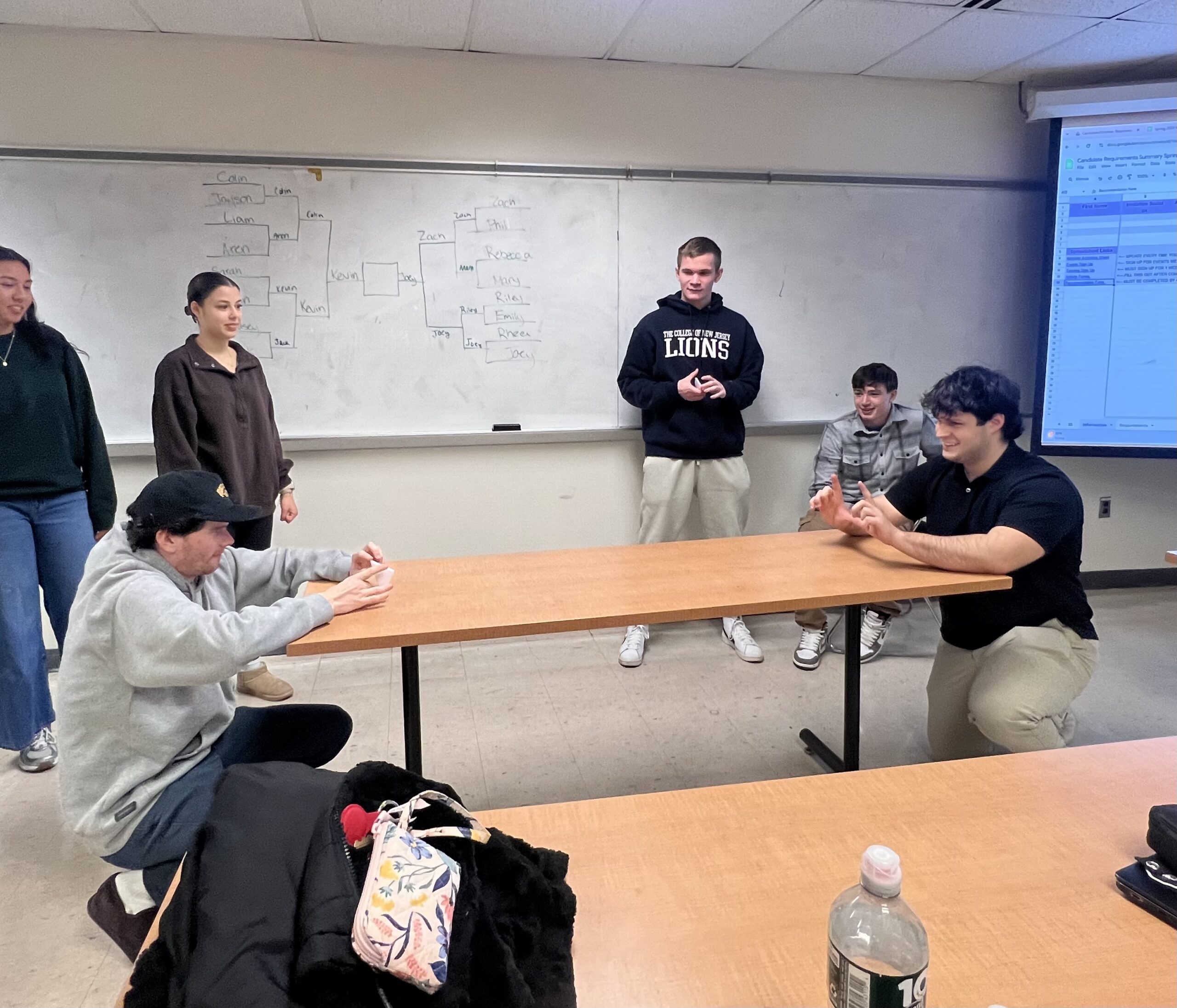 Two male students kneeling at each end of a table playing table football. One students is kicking a paper triangle (ball) with his fingers and another is making a goal space with his. Several students stand in the background observing.