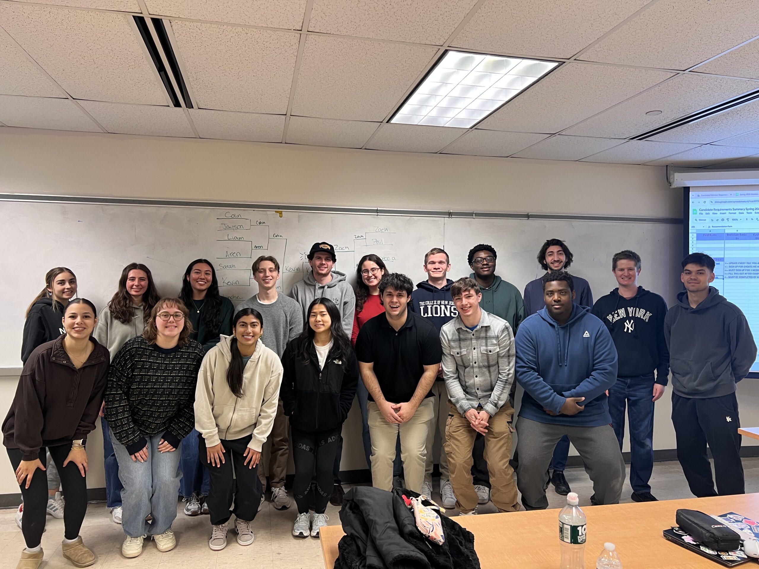 A group of students standing in two rows in front of a whiteboard.