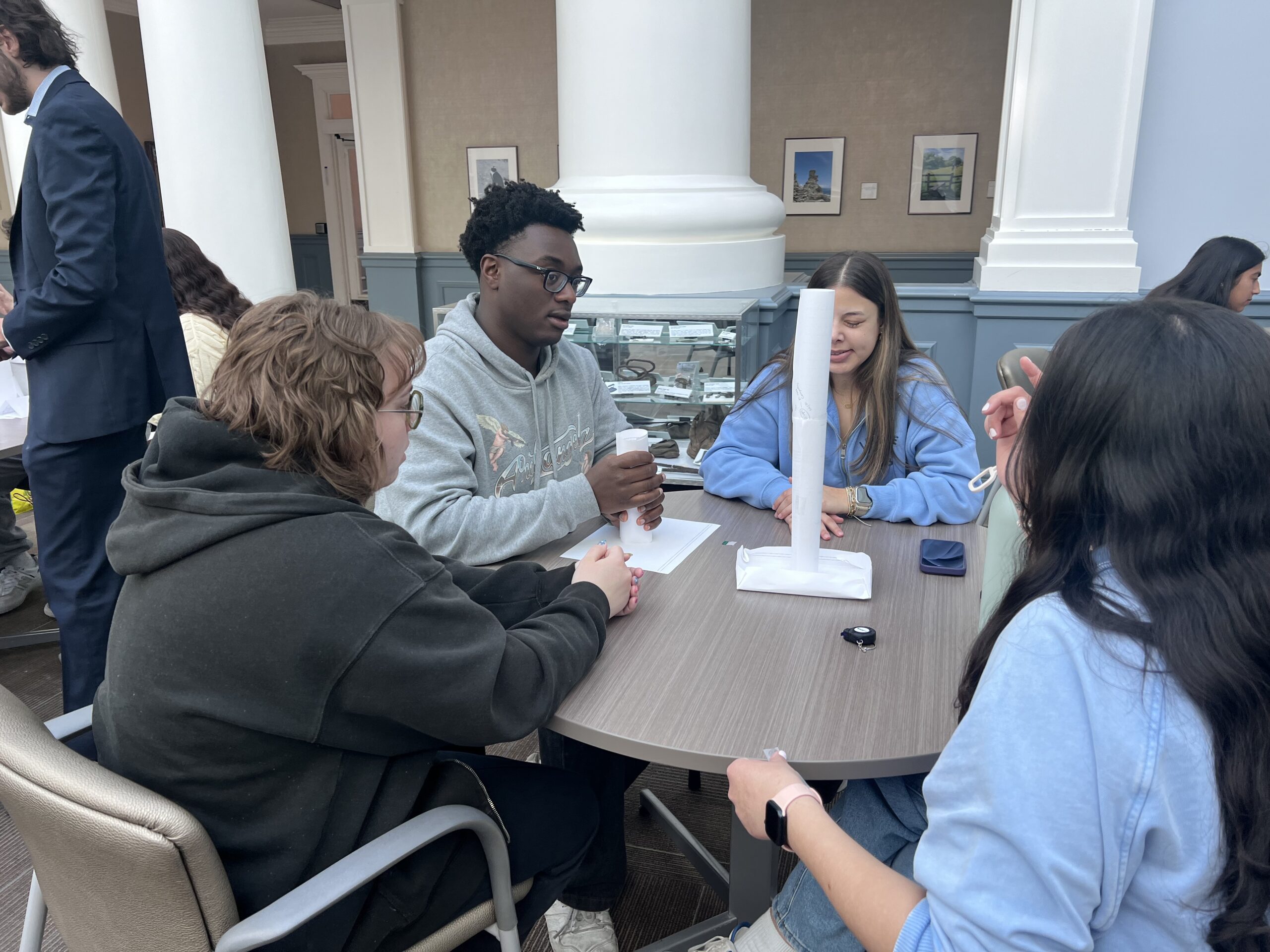 Four students sit around a table constructing a paper tower.