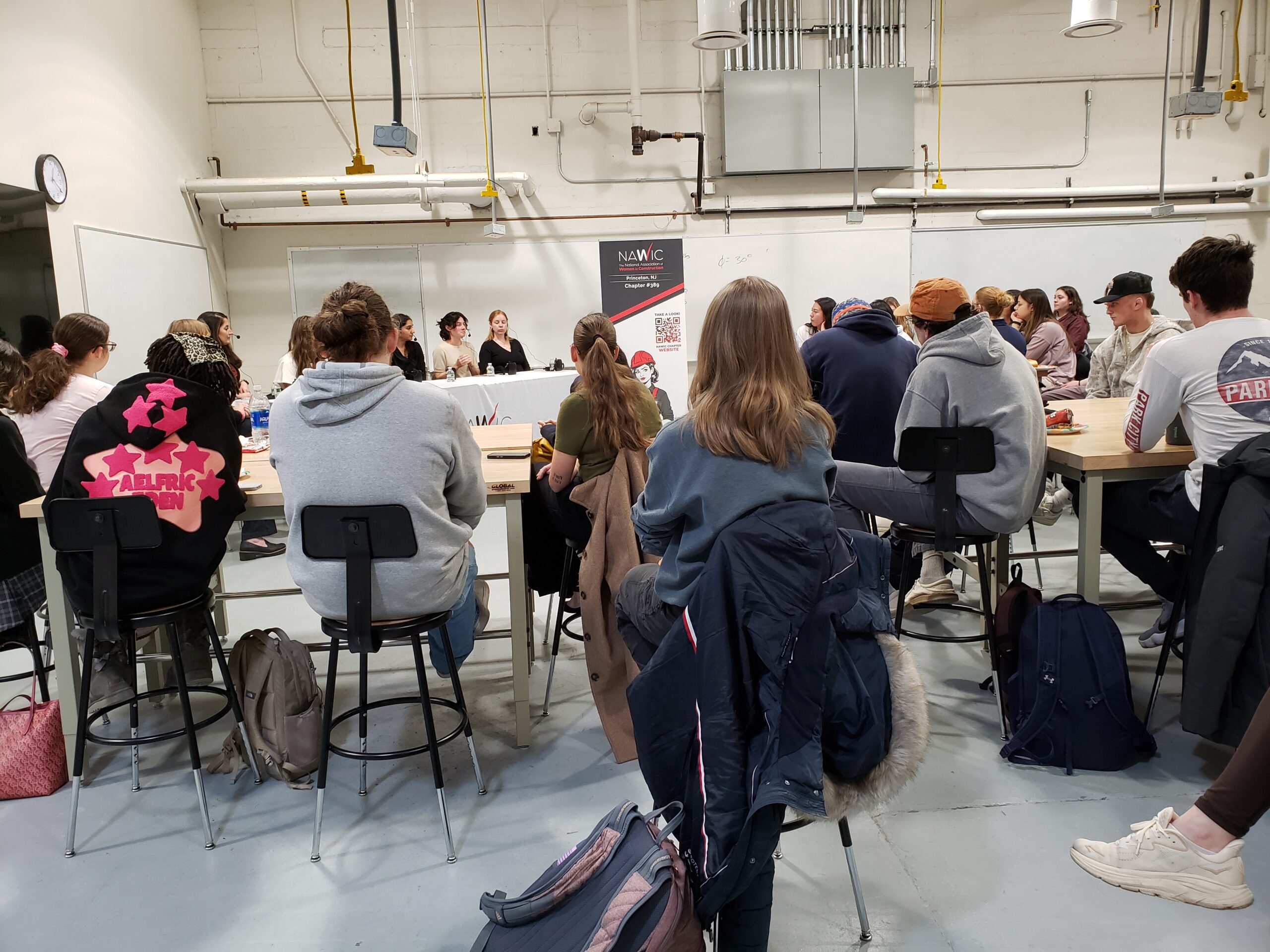 A large group of students sitting at tables s photographed from behind. A table with panelists can be seen in front of the group.