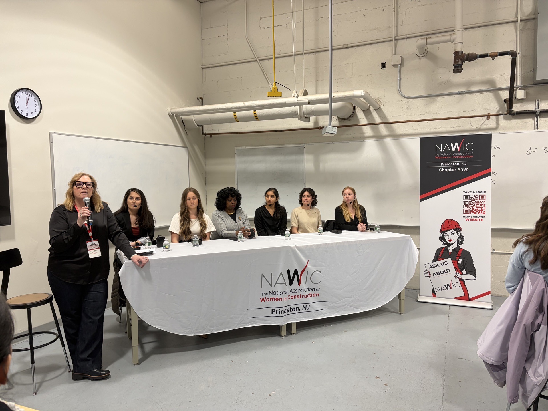 One woman standing next to a long table that seats six other women.