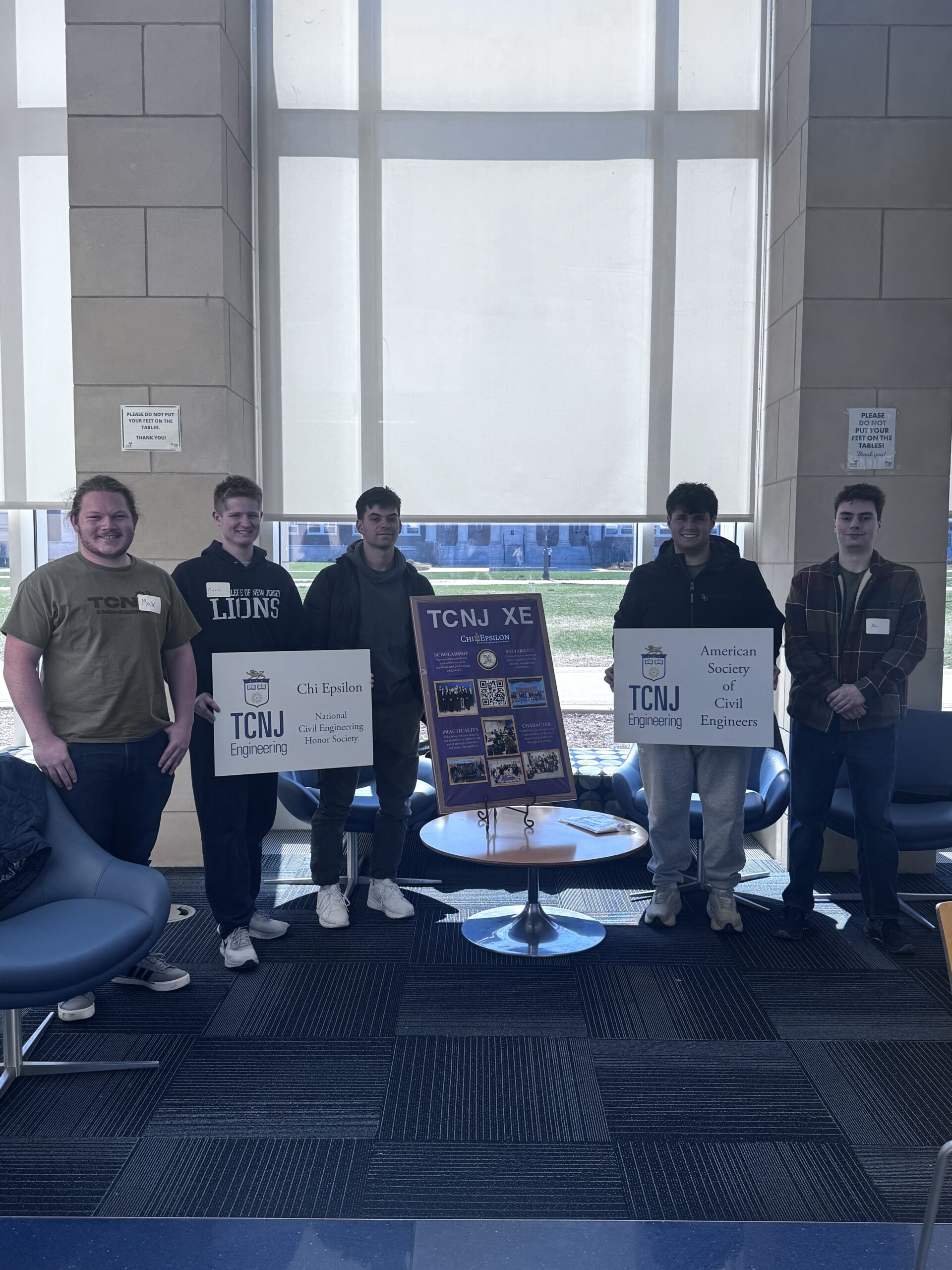 Five male students standing in front of a large window. Three stand on the left of a display board that outlines TCNJ XE activities and two on the right side. One student on the left and one student on the right hold smaller signs in their hands.