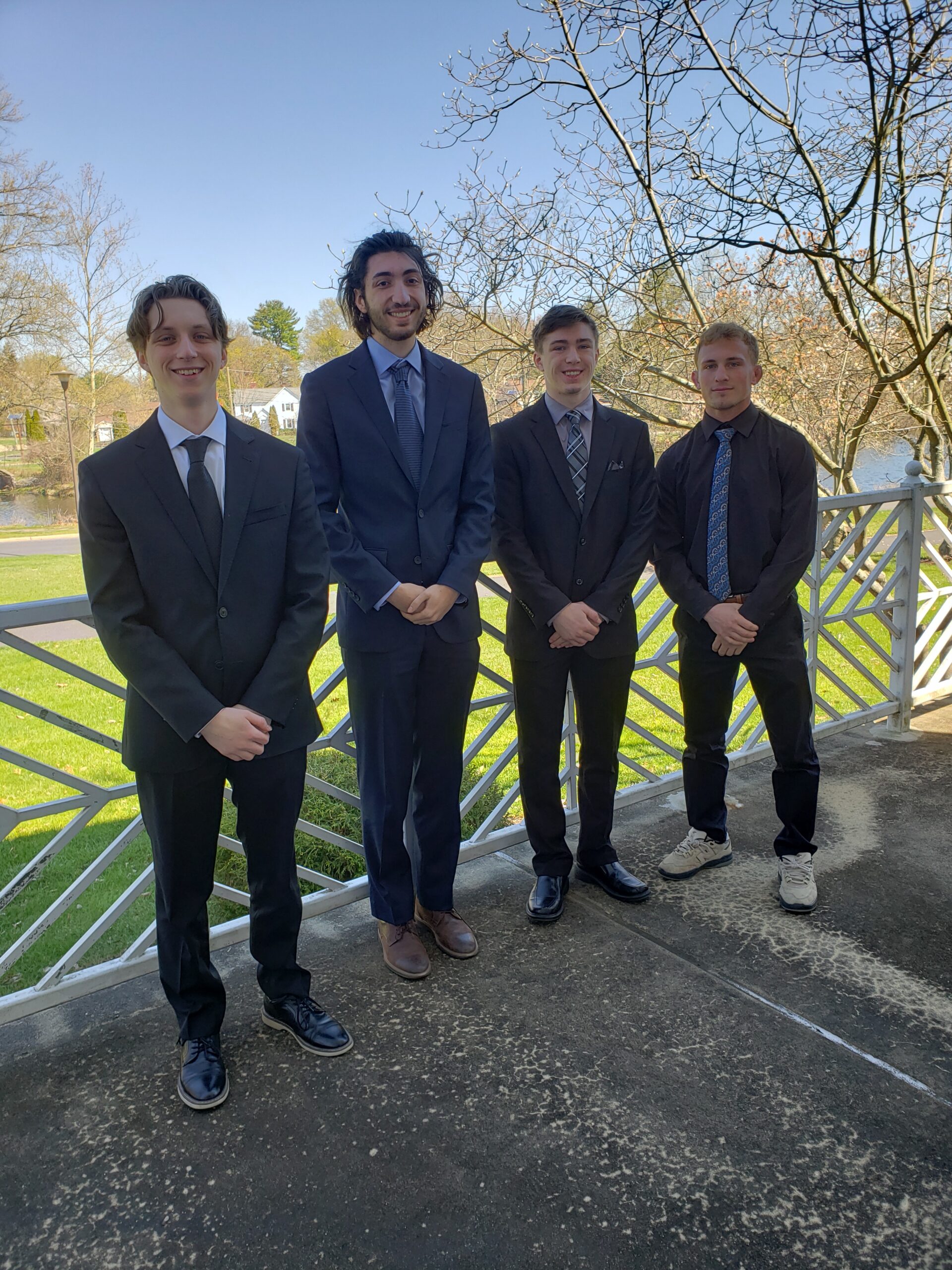 Four male students in suits and ties stand on a patio