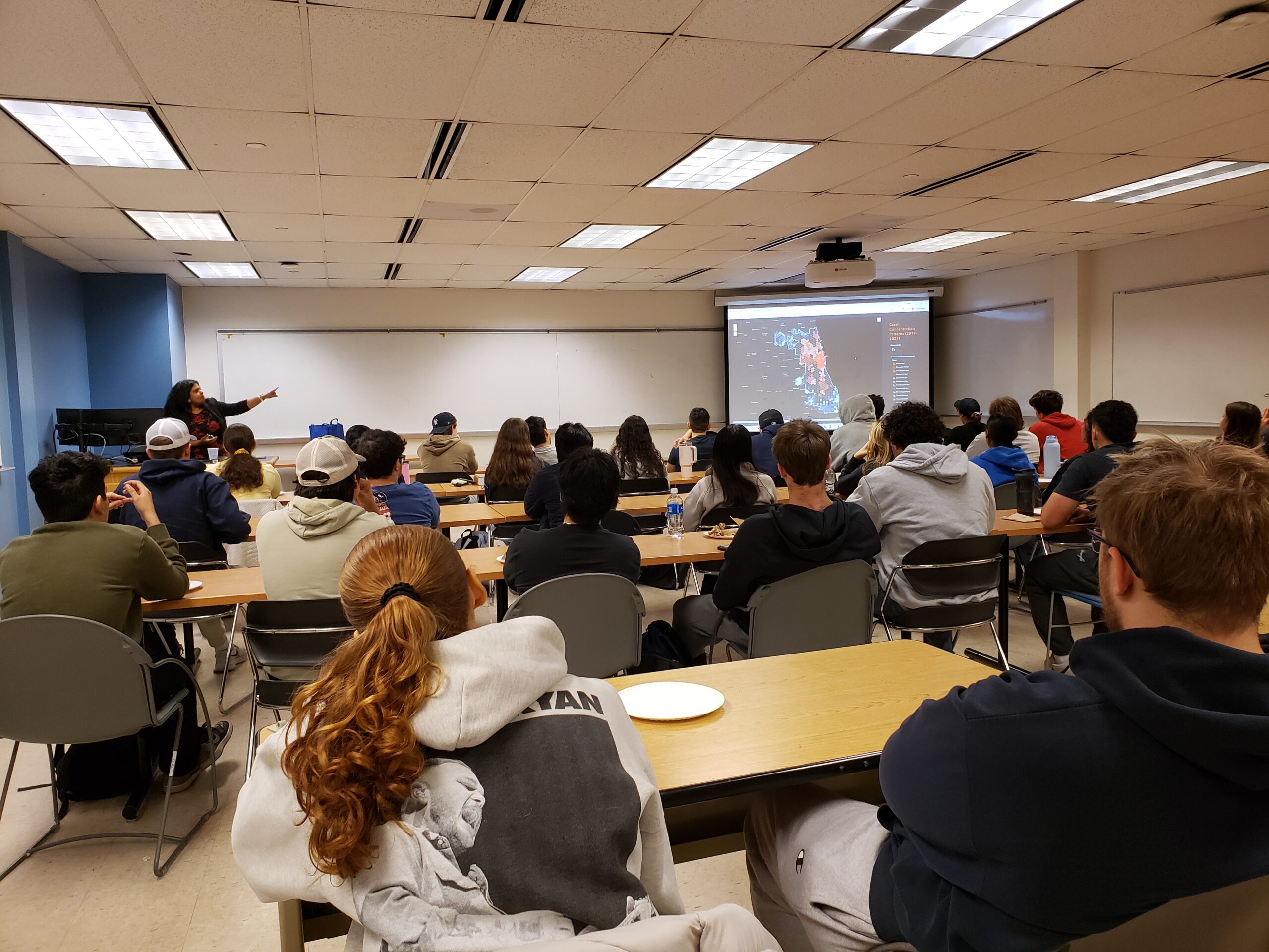 A group of students sitting at desks in a classroom (view from behind) with the screen in front of them