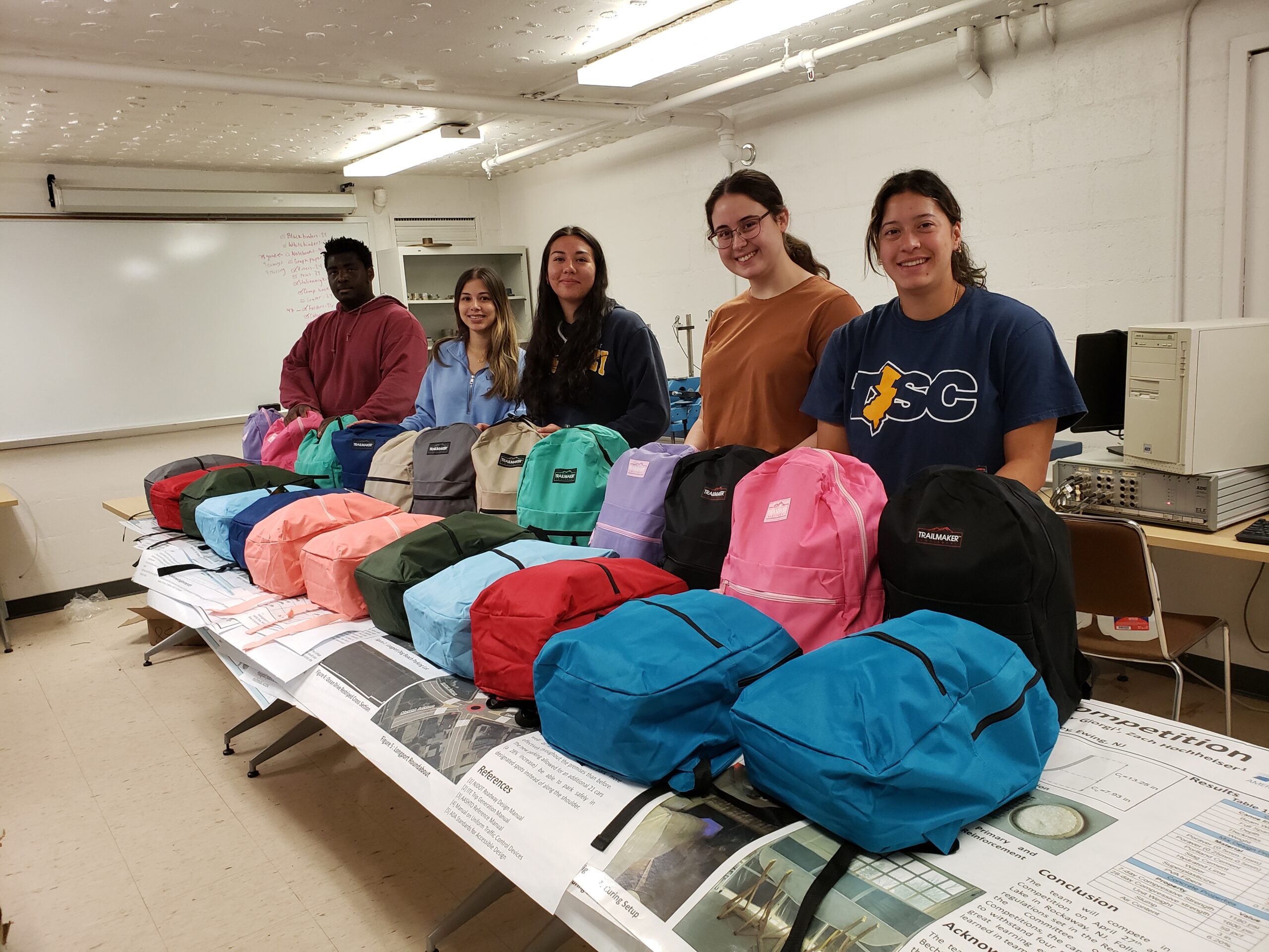 Five students stand behind a table that holds 24 school bags filled with supplies
