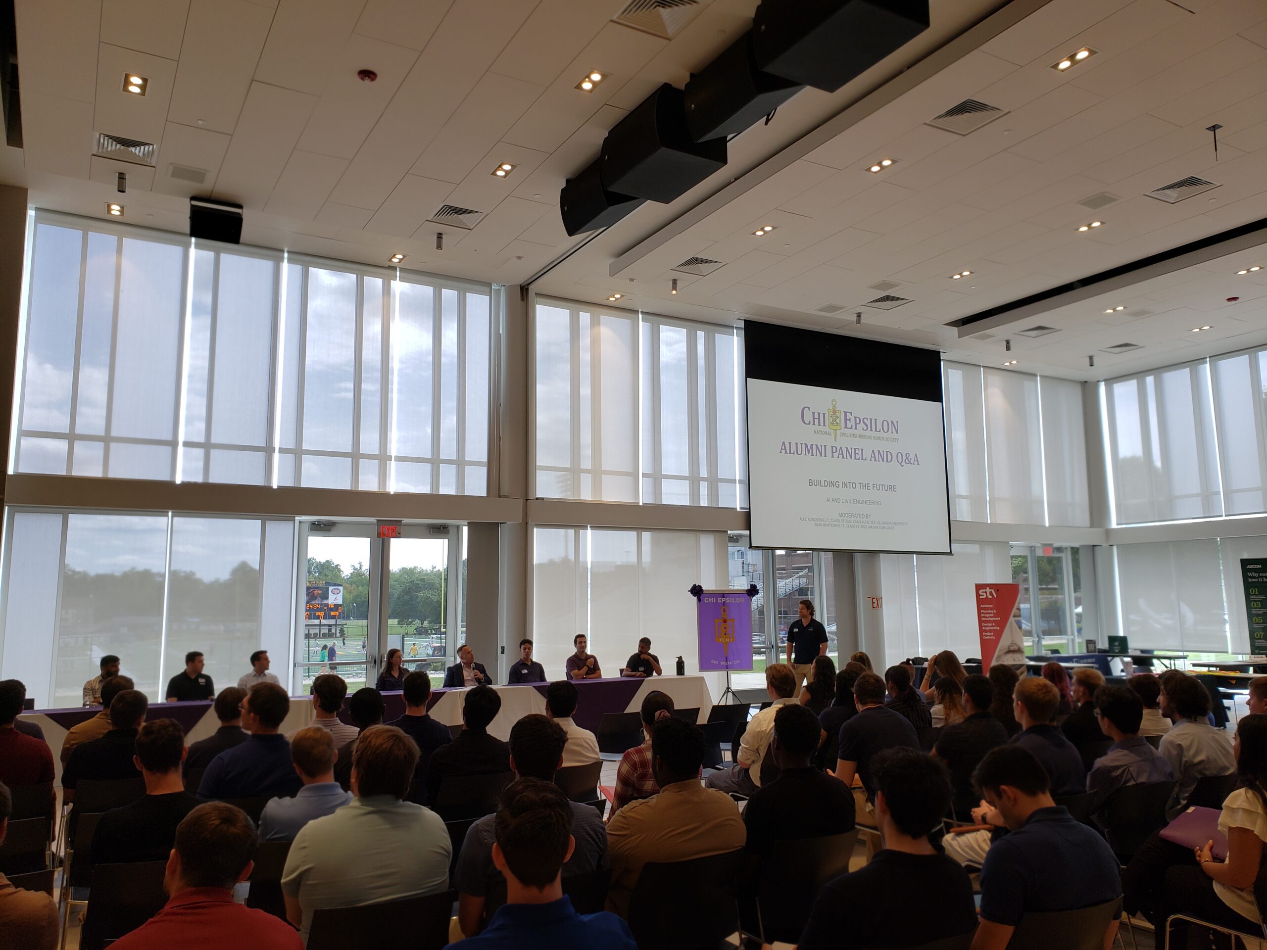 Panel room setup with 8 people facing the room of students. There is a screen in the background that displays the name of the event.