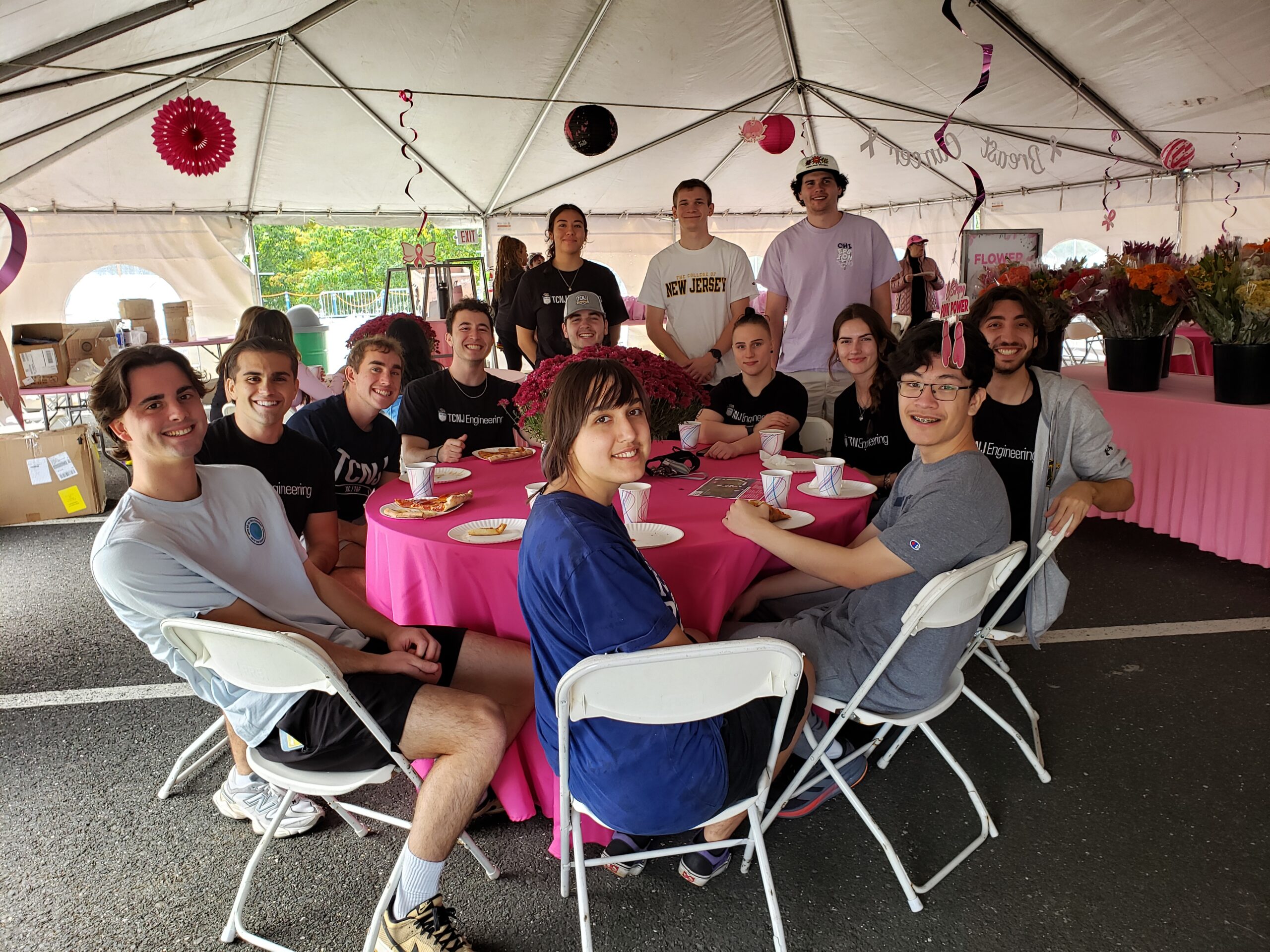 A large group of students sit around a round table with three students standing in the background.