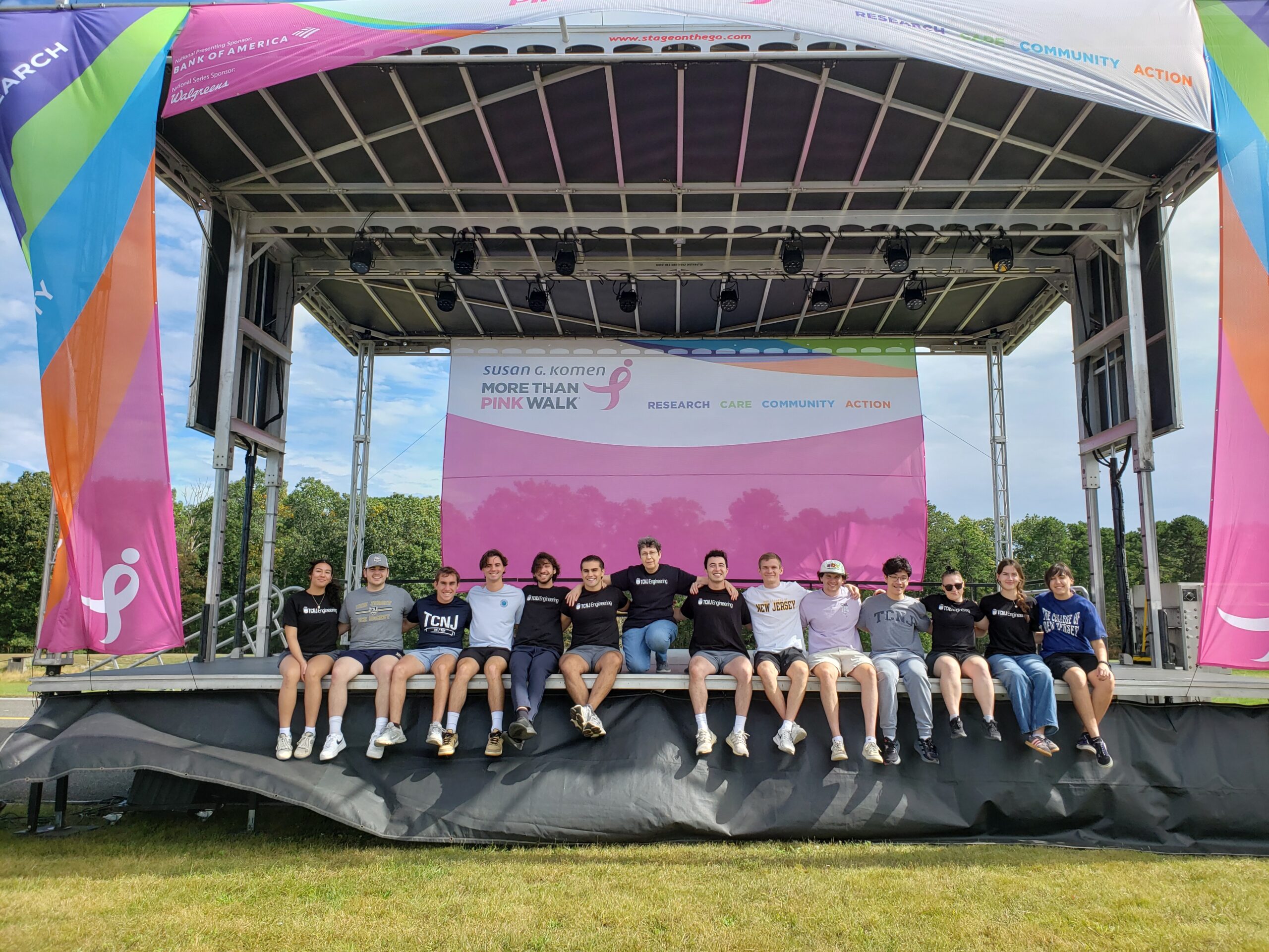 A group of students sit at the edge of a large event stage with their professor crouching in the middle of the row. Everyone has their arms around each other's shoulders.