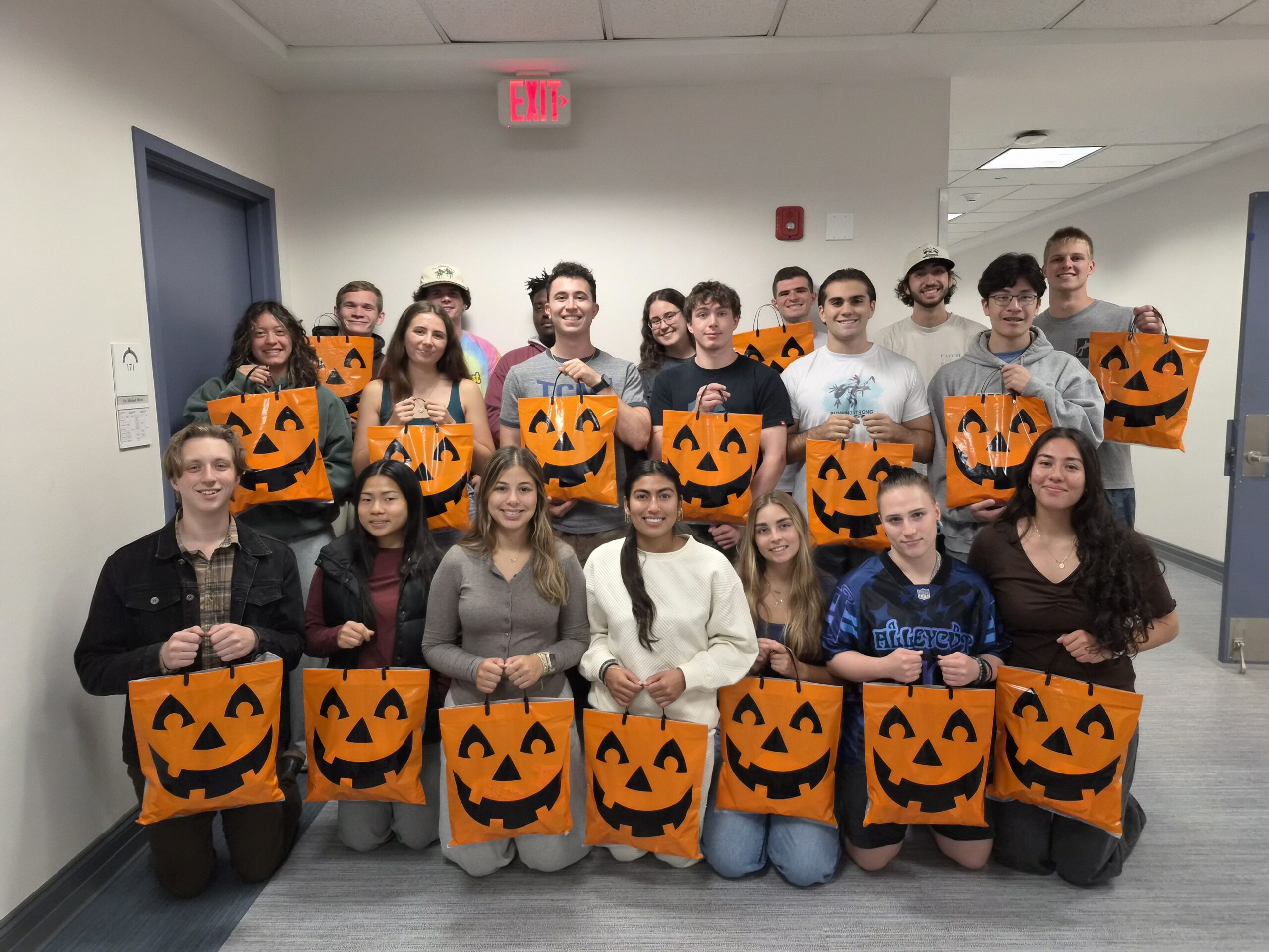 A large group of students standing in three rows and holding orange pumpkin Halloween bags. The front row of students is kneeling and the second and third row are standing behind them.