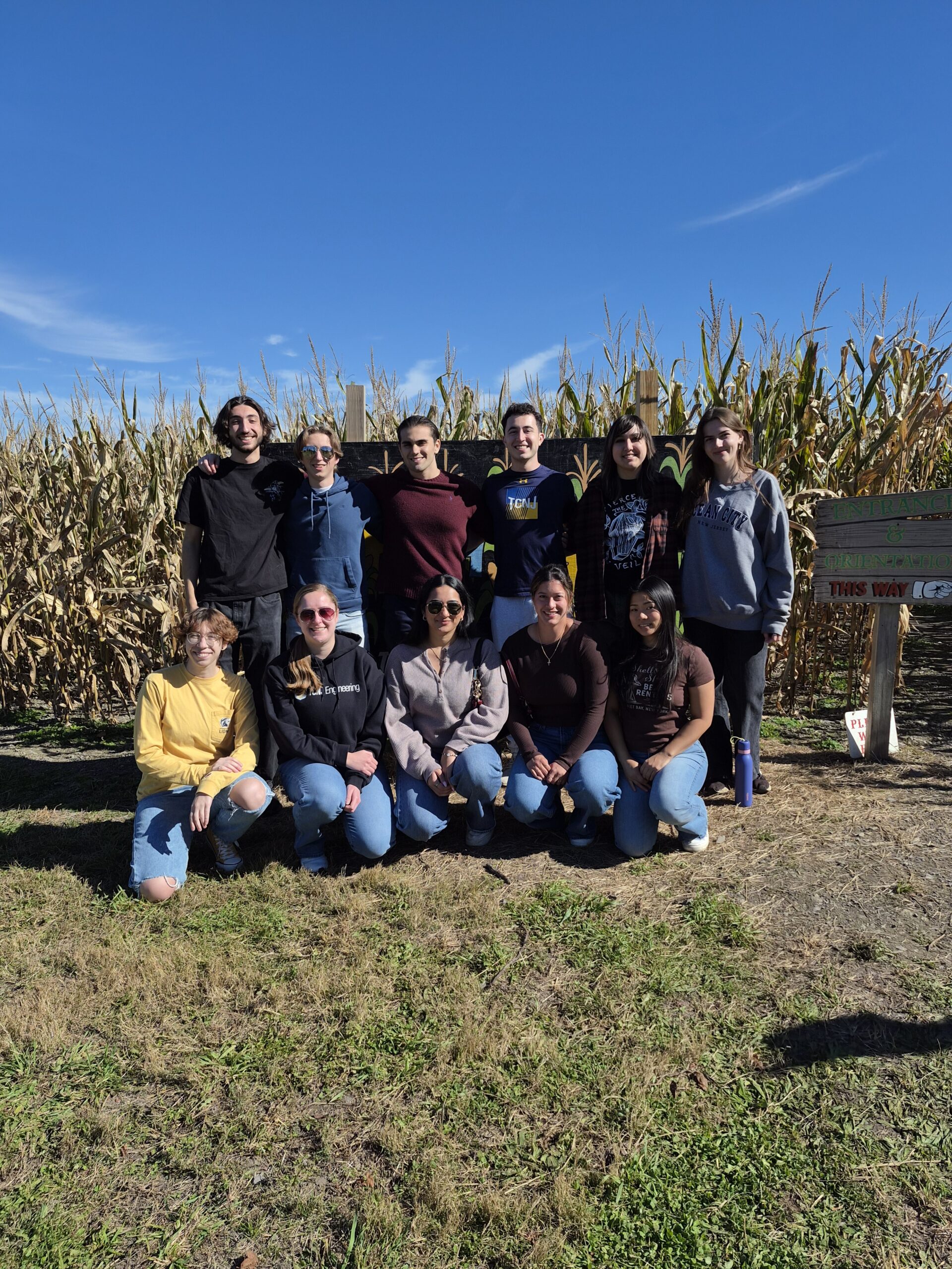 A group of young people stands in front of the corn maze. Back row with 6 students is standing and the front row with 5 student is squatting.
