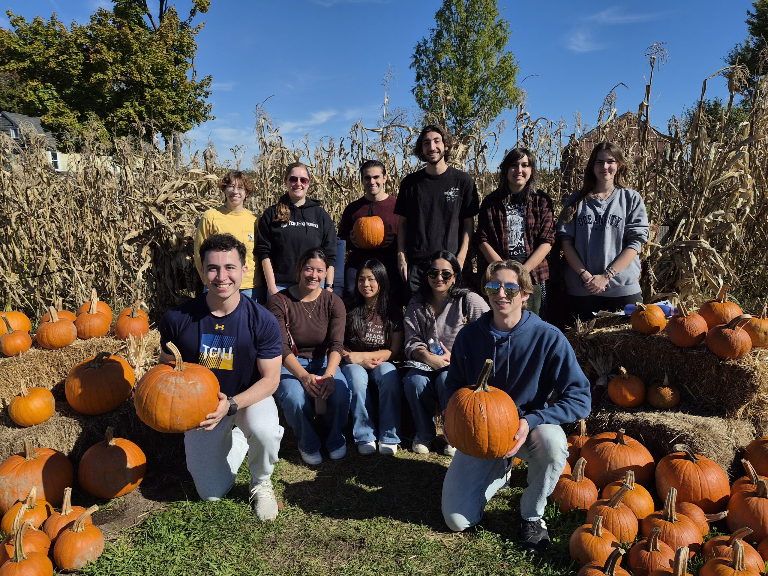 A group of young people are in front of a corn field. Back row stands and the frong row is either sitting on hay bales or squatting. Two students on each end of the front row hold large pumpkins. One person in the back row is also holding a pumpkin.