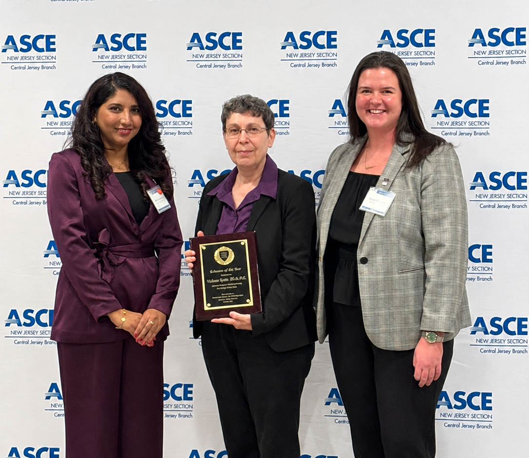 Three women stand in front of a white backdrop that reads ASCE multiple times. The woman in the middle wears a black suit and a purple shirt and holds the award.