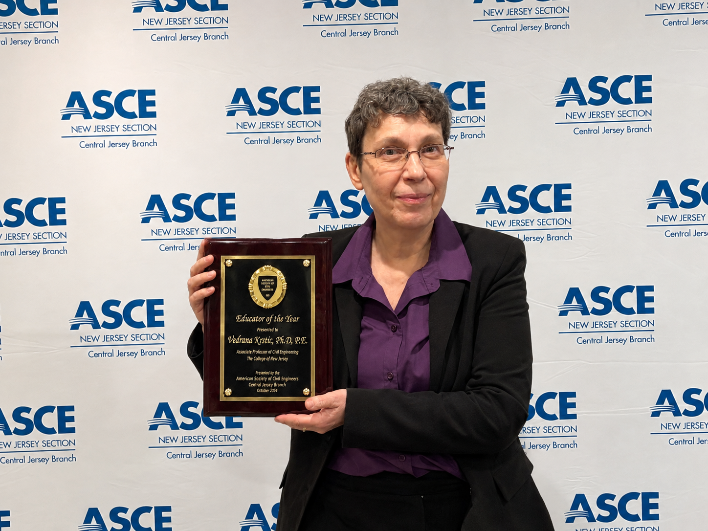 A middle-aged woman in black suit and purple shirt with short hair and glasses holds an award plaque. 